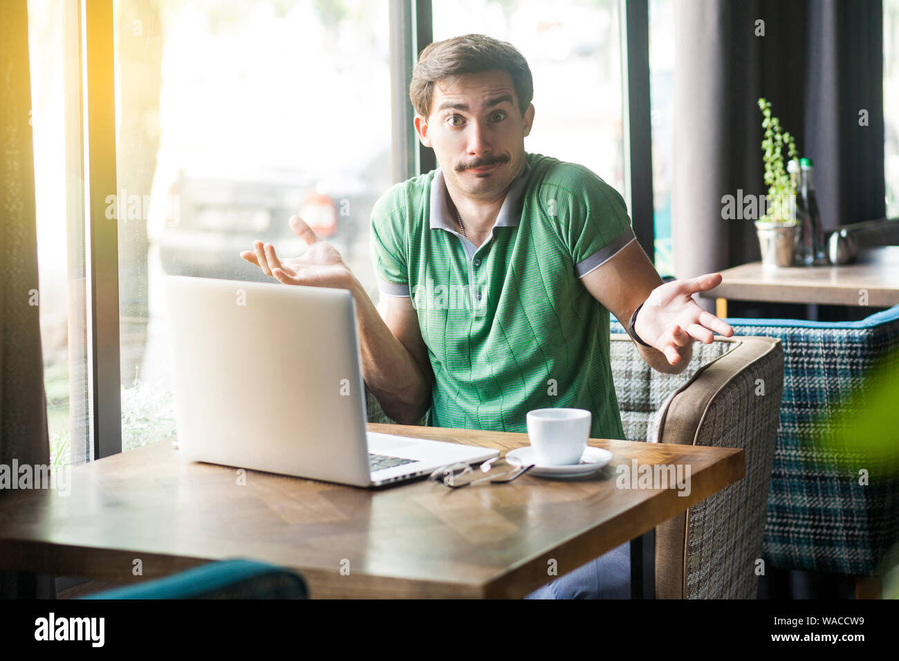 Non lo so! Giovane imprenditore confuso in verde t-shirt seduta con laptop, guardando la telecamera con i bracci sollevati e perplesso. business e freelancin Foto Stock