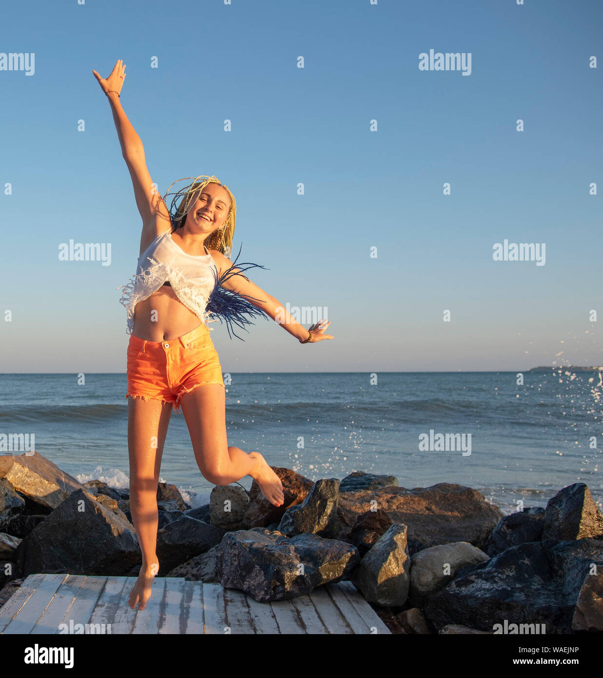 Ragazza senegalese con pig-tail salta sul pontile in legno sul mare. Le onde sono di colpire le rocce e gli spray è volare in direzioni diverse. V Foto Stock