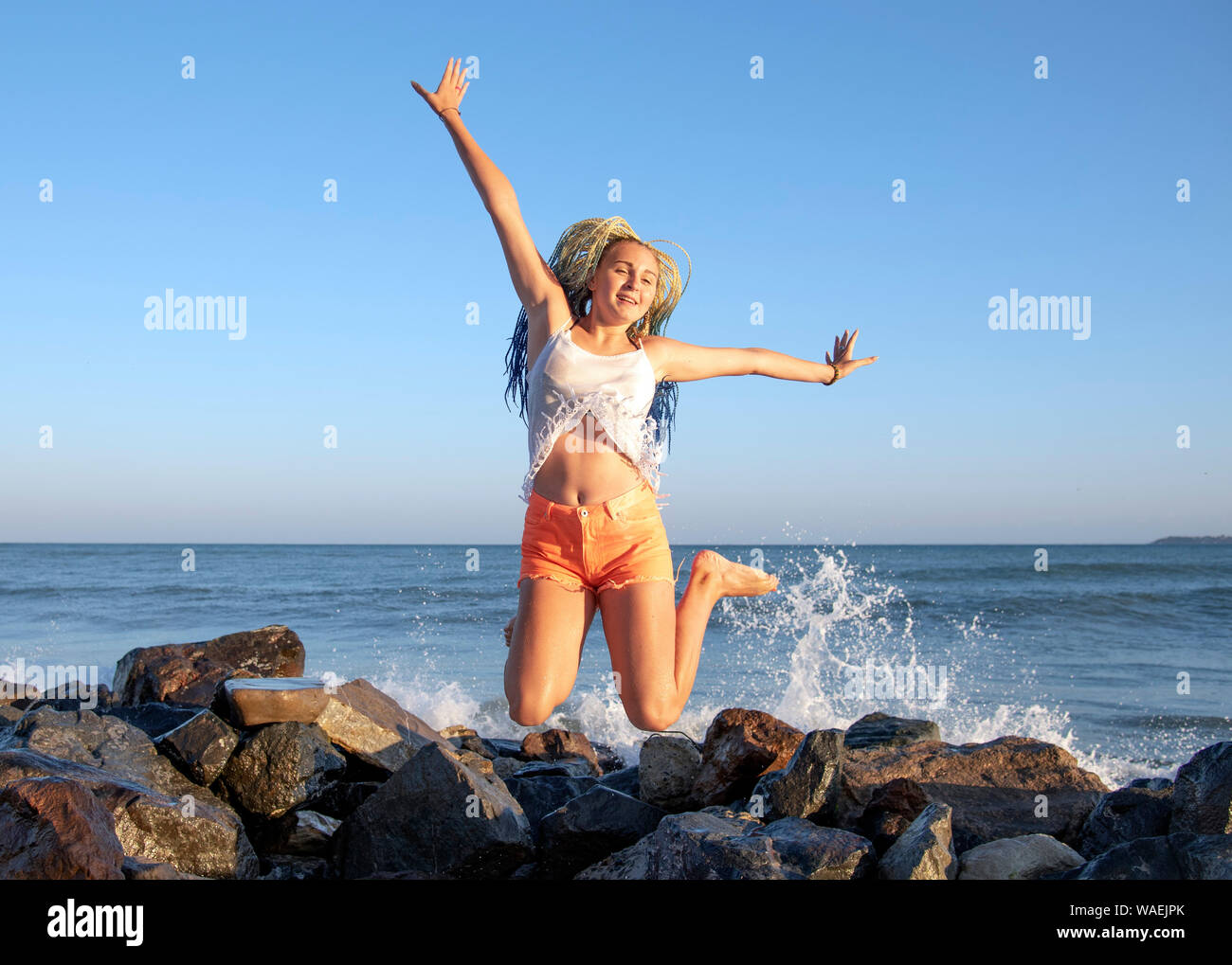 Ragazza senegalese con pig-tail salta sul pontile in legno sul mare. Le onde sono di colpire le rocce e gli spray è volare in direzioni diverse. V Foto Stock