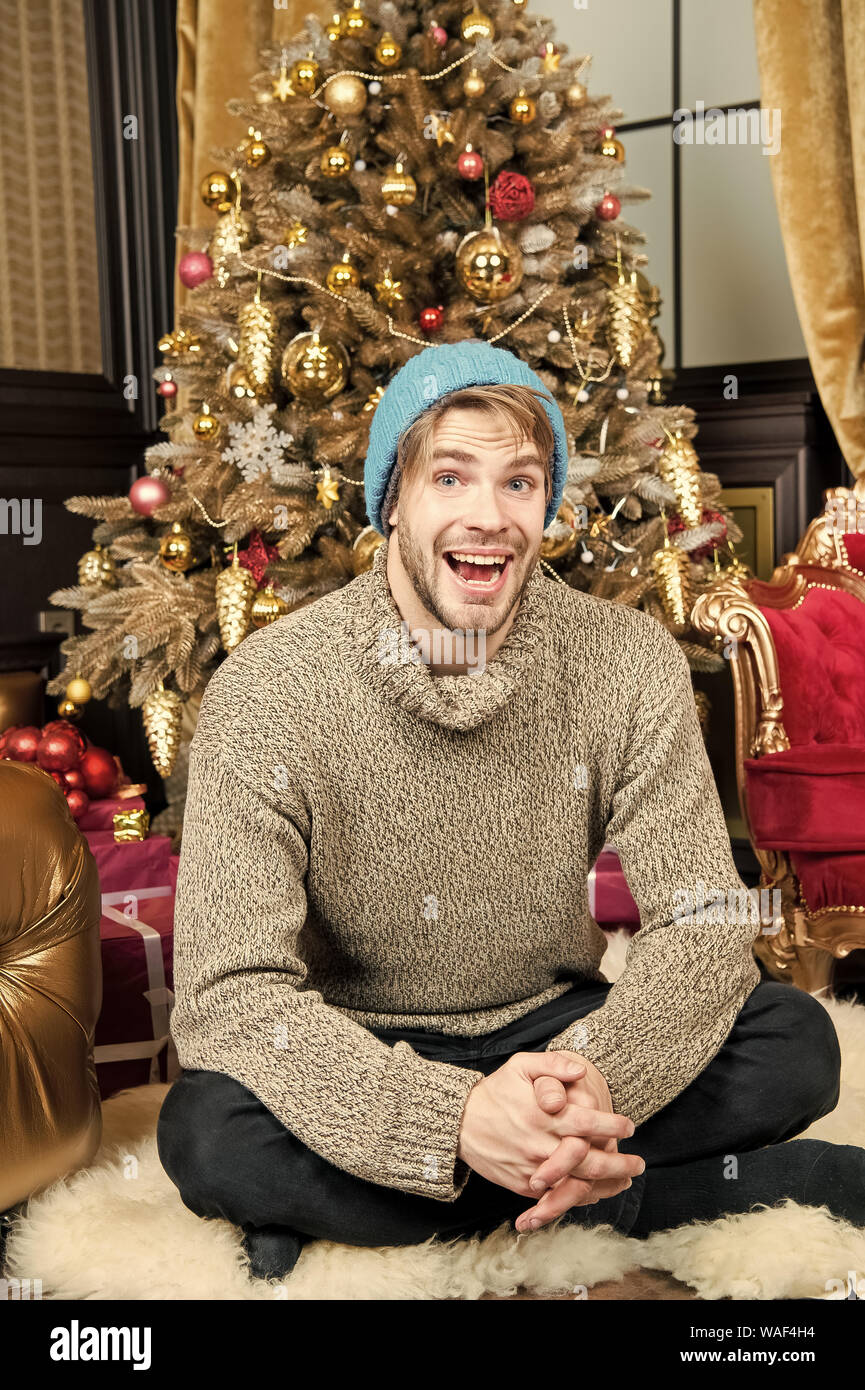 Macho sorriso sul tappeto peloso a albero di natale in camera. Uomo seduto  in un caldo cappello, maglione ad albero di Natale. Vacanze a preparazione  e celebrazione. Felice anno nuovo, eve, partito.