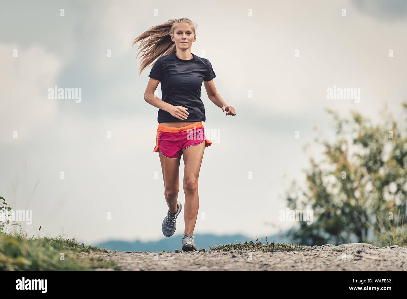 Bella bionda atleta corre su strada sterrata in collina Foto Stock