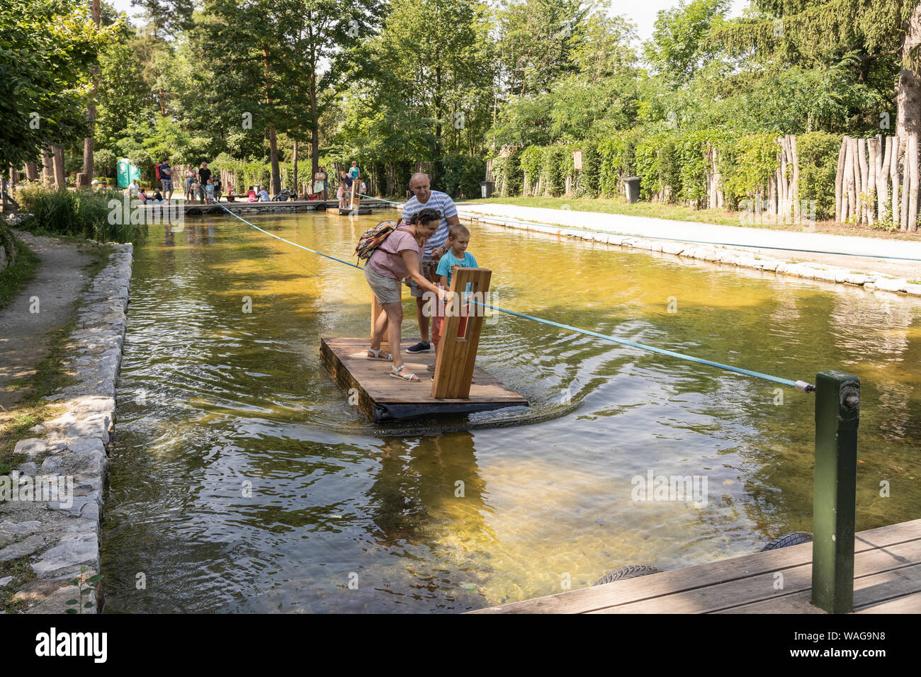 Visitatori godendo una giornata fuori a Kittenberger Erlebnisgärten tirando stessi attraverso un laghetto con un traghetto via cavo Foto Stock