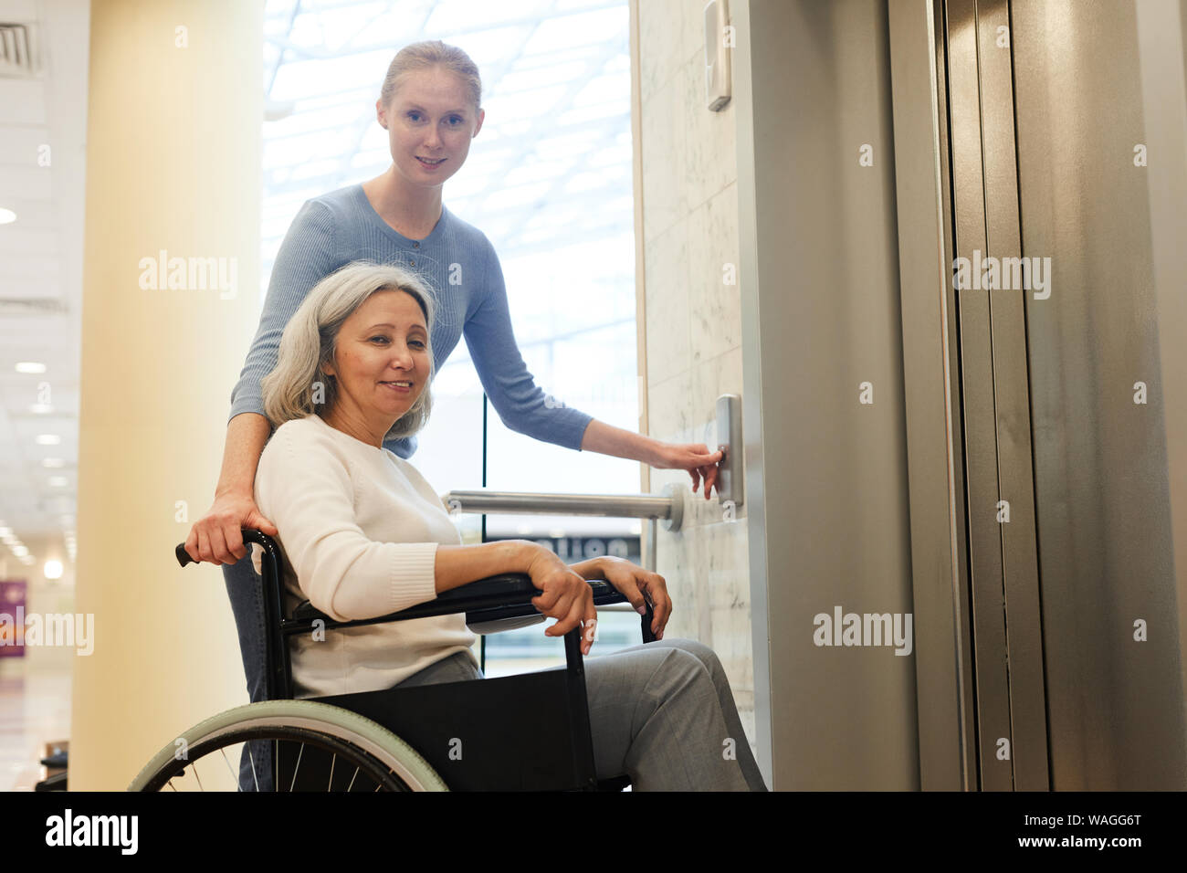 Ritratto di giovane infermiere senior con paziente in carrozzella sorridente in telecamera durante l'attesa per l'ascensore all'ospedale Foto Stock