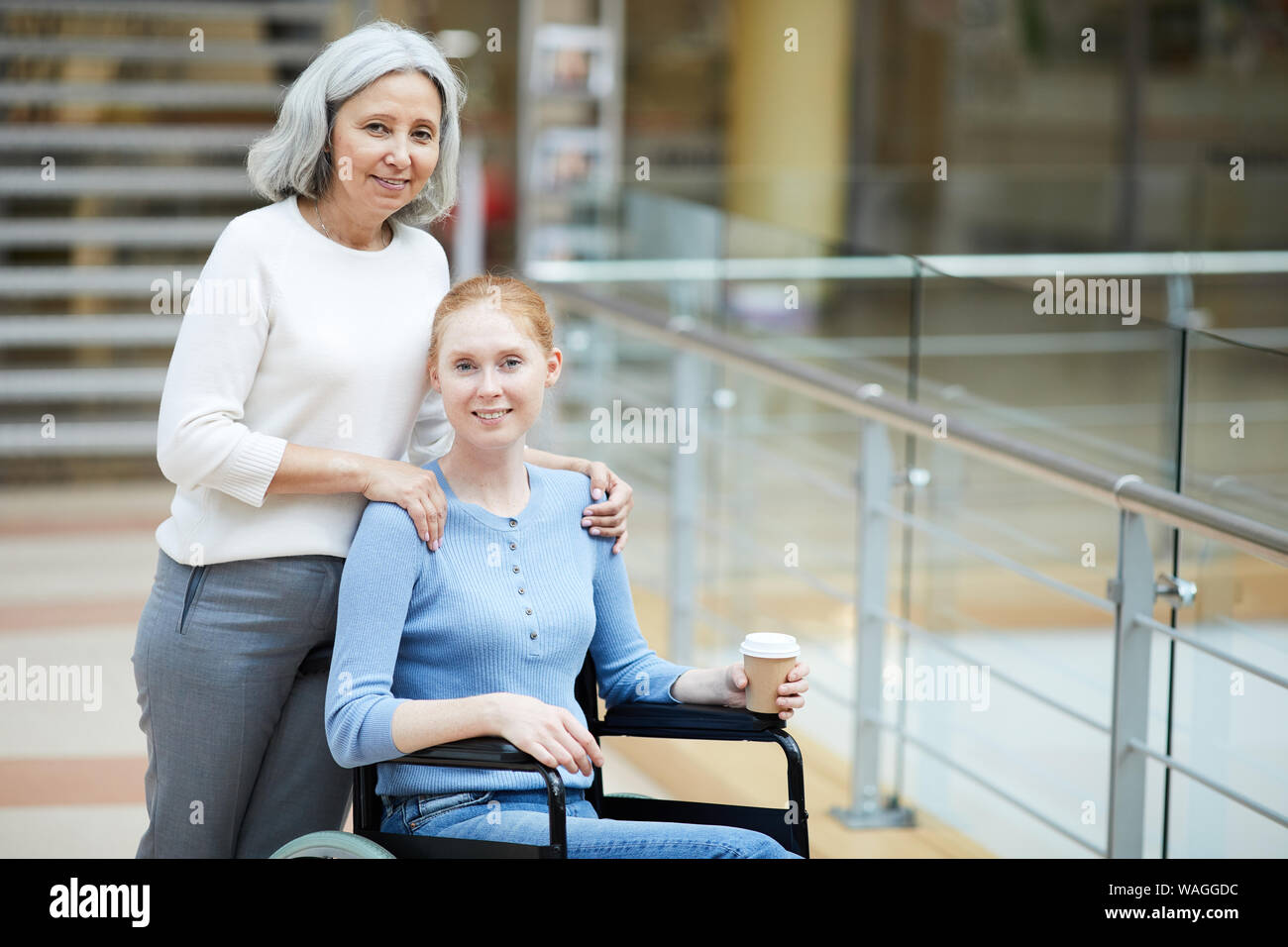 Senior madre abbracciando la sua figlia di disabili in carrozzella ed essi sorridente in telecamera nel moderno centro commerciale Foto Stock