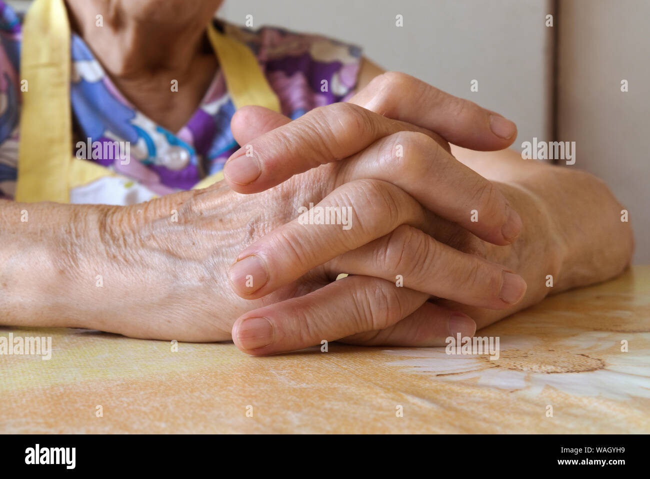 Donna Senior in appoggio con le mani incrociate su un tavolo da cucina. Old Lady indossando pinafore colorati, il fuoco selettivo, illuminazione naturale, vista laterale. Foto Stock