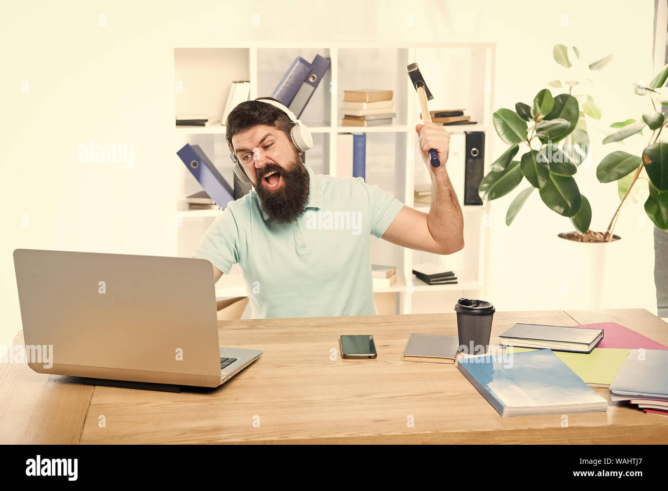 Odio di ufficio di routine. Uomo Barbuto guy ufficio cuffie swing martello sul computer. Connessione a internet lenta. Software obsoleto. Computer lag. Ragioni per computer in ritardo. Come fissare il lento sistema di ritardo. Foto Stock
