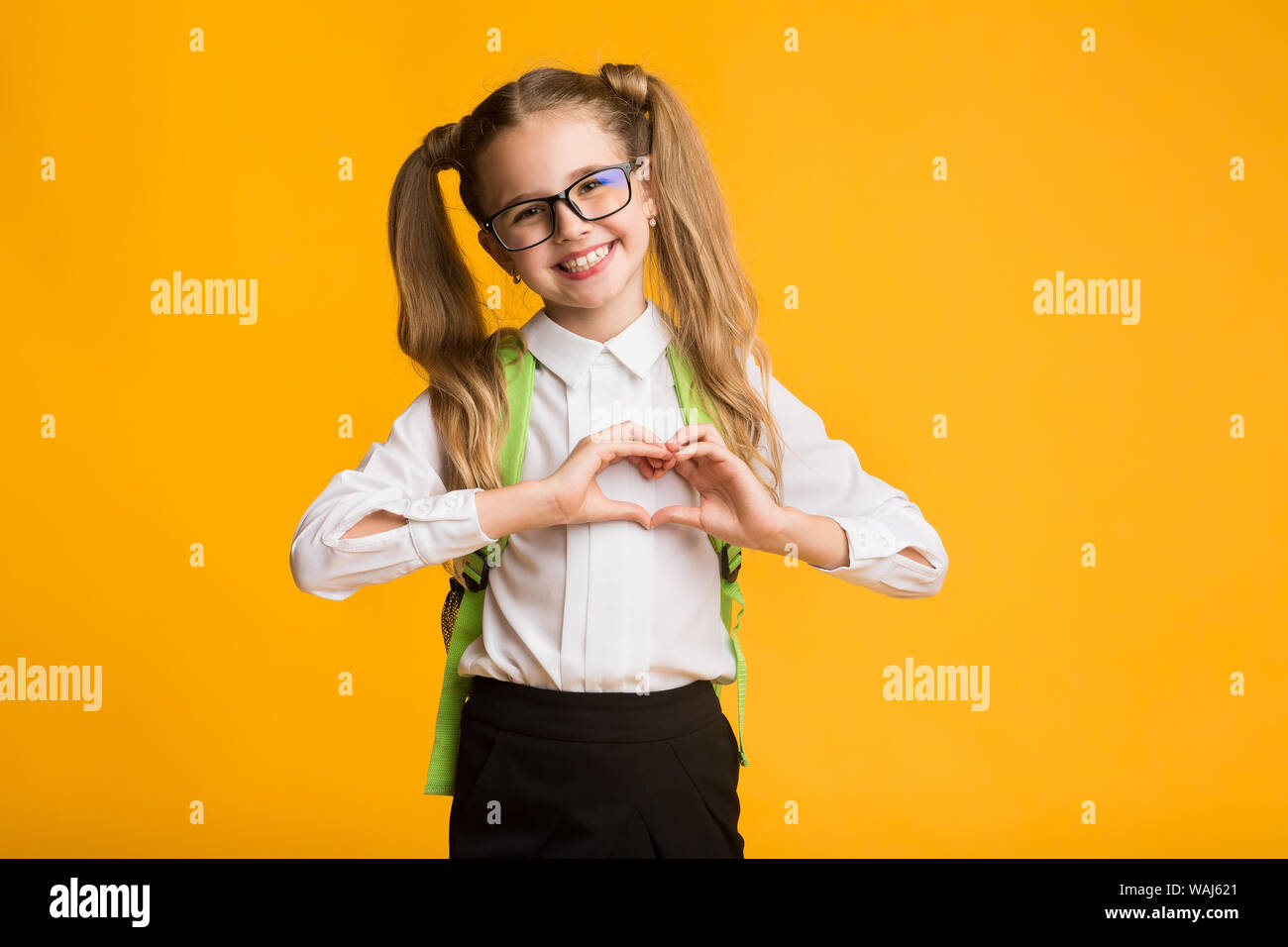 Little Schoolgirl sorridente che mostra il cuore con le mani su sfondo giallo Foto Stock