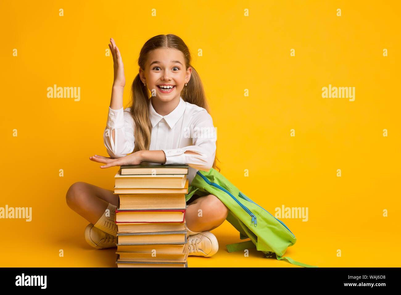 Clever Schoolgirl alzando la mano seduto alla pila di libri, sfondo giallo Foto Stock