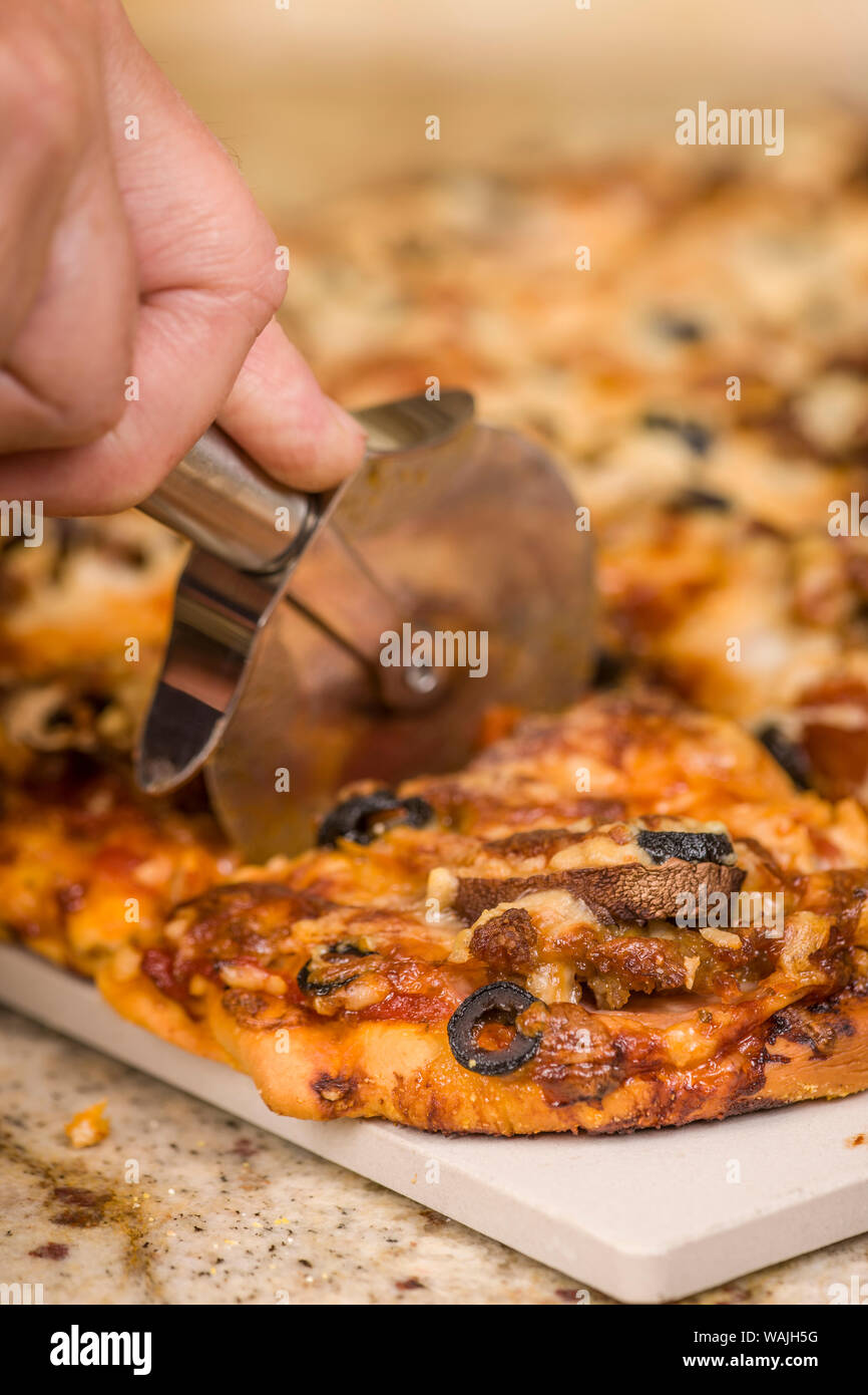L'uomo il taglio di un pezzo di carne fatti in casa e veggie pizza con condimenti di salsiccia, salsiccia per pizza, Canadian bacon, olive nere, funghi, mozzarella, parmigiano e pecorino romano. (MR) Foto Stock