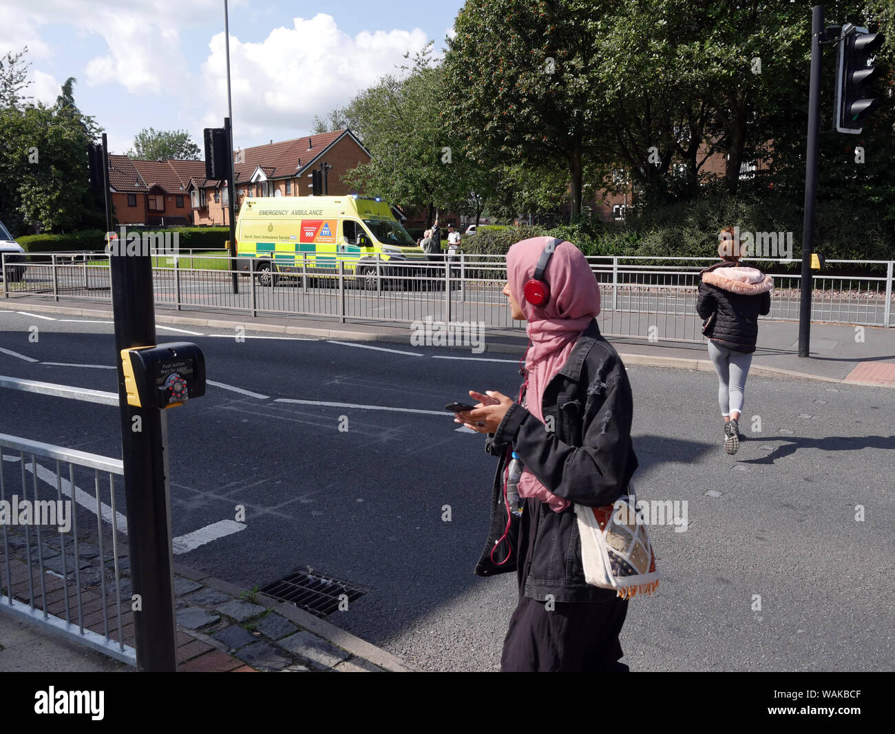 Femmina con smartphone & auricolari guarda la scena dell'incidente su un attraversamento pedonale sulla strada a doppia carreggiata, modo Topp, Bolton, England Regno Unito. fotografia DON TONGE Foto Stock