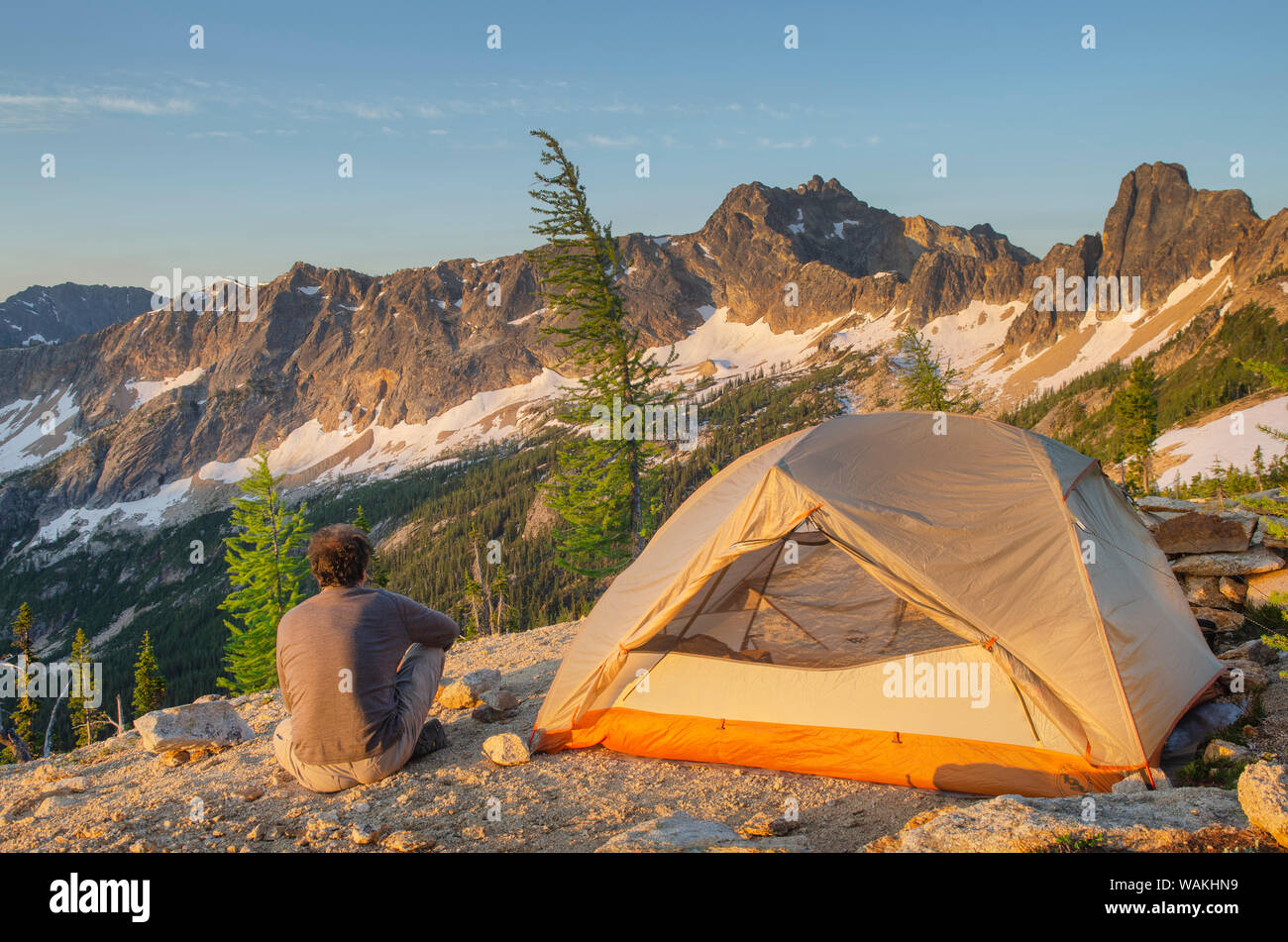 Uomo adulto allo spuntar del giorno seduto accanto a backpacking tenda e guardando a vista del picco spietato sulla cresta sopra Tagliagole Pass, vicino Pacific Crest Trail. North Cascades, nello Stato di Washington Foto Stock