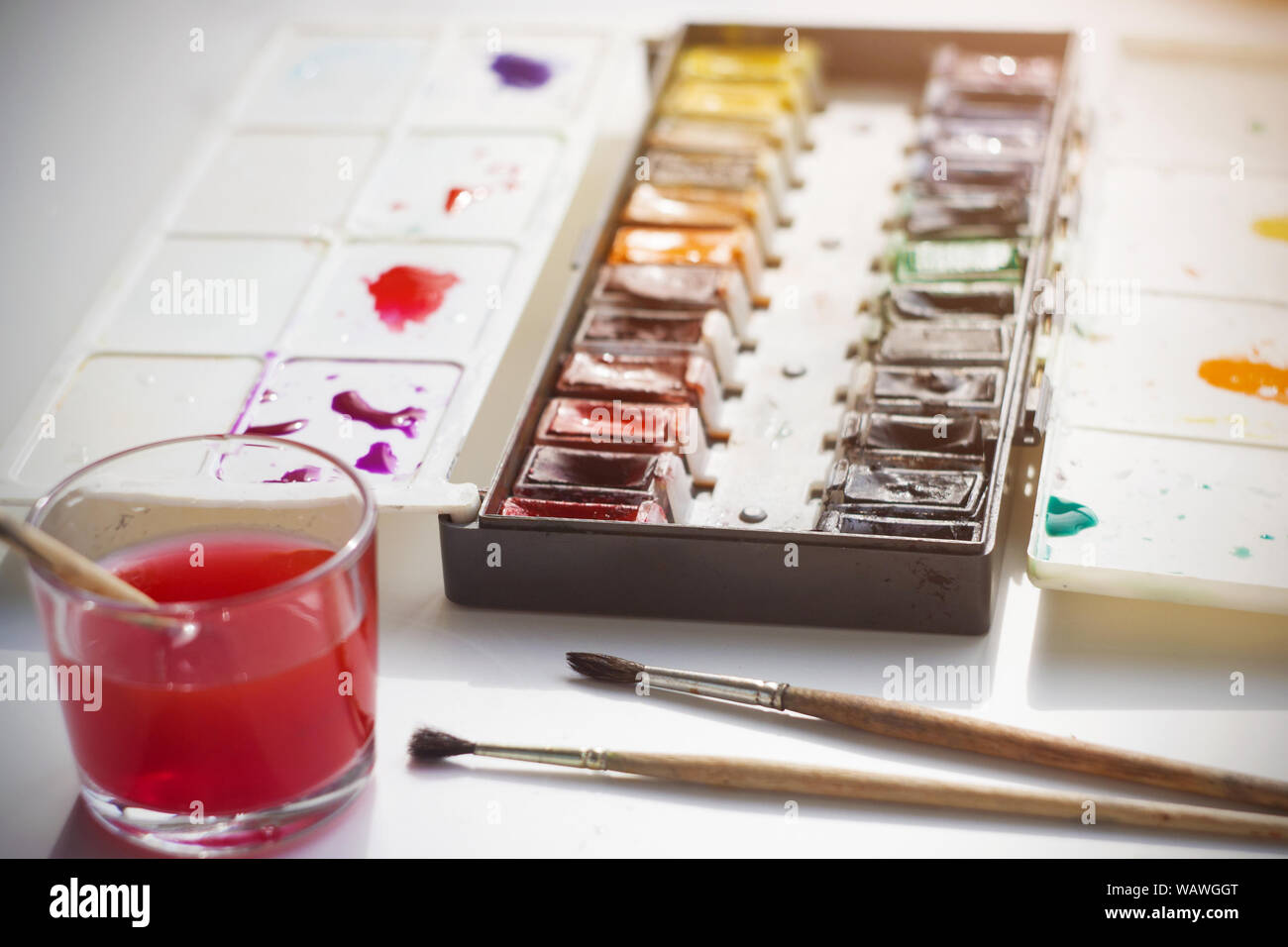Su un tavolo bianco, illuminato dalla luce del sole, vi sono pitture ad acquerello in una scatola di plastica, spazzole per disegno e un bicchiere di acqua per il lavaggio della spazzola Foto Stock