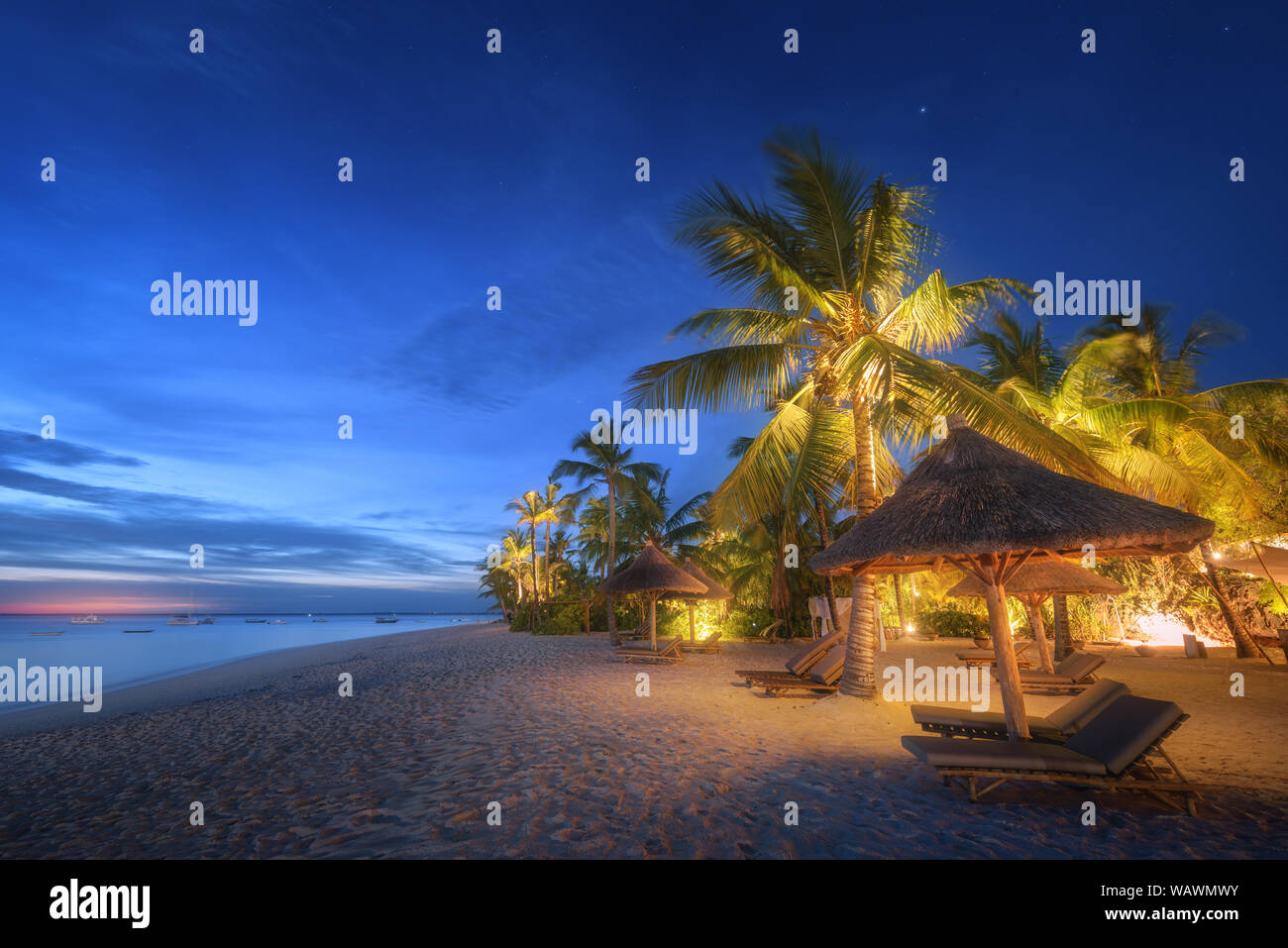 Spiaggia di sabbia con palme e sedie a sdraio sotto gli ombrelloni in blu ora Foto Stock