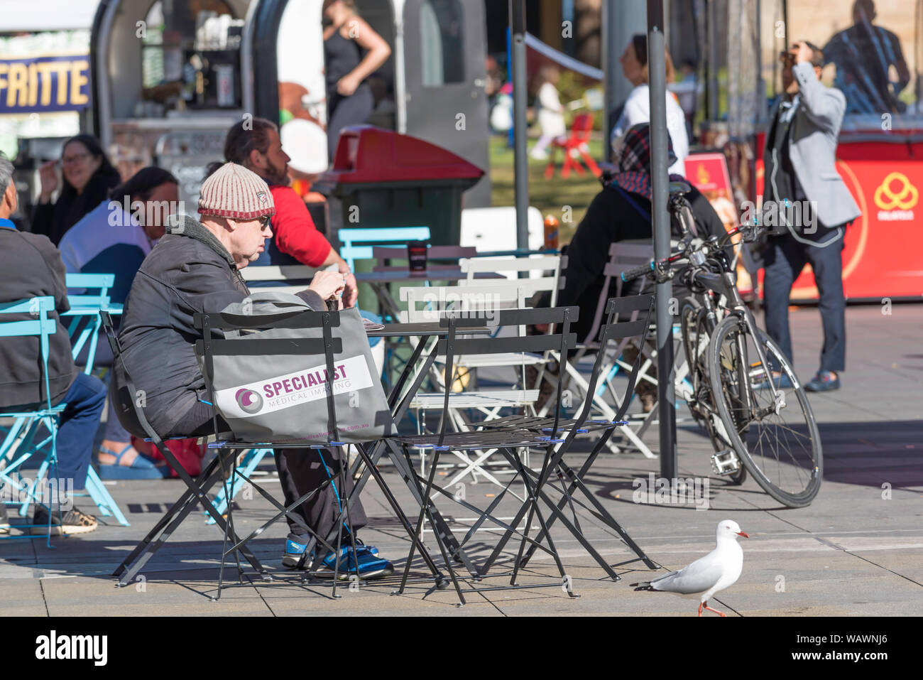 Un vecchio uomo bianco che indossa un beanie siede sul peso di luce posti a sedere esterni in Centenary Square, Parramatta, Australia in una giornata di sole in inverno Foto Stock