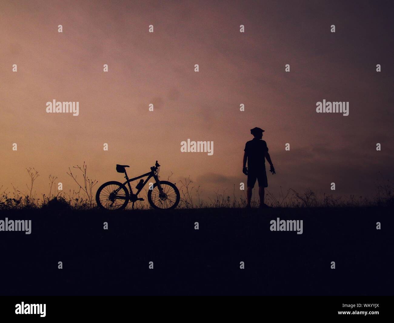 Silhouette di un maschio in piedi vicino a una mountain bike su una collina con un cielo viola sullo sfondo Foto Stock