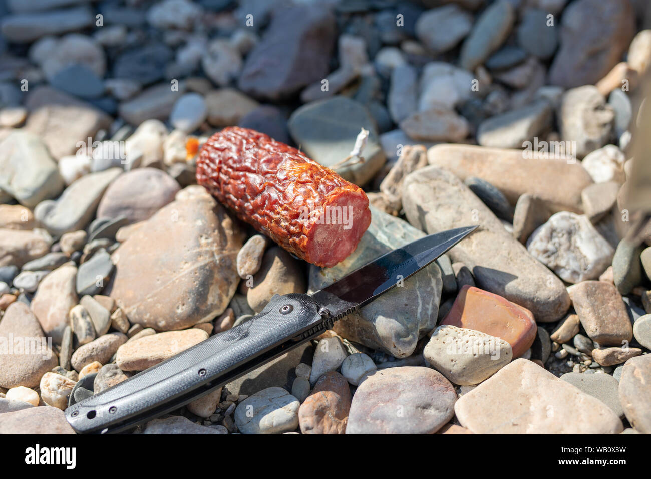 Affumicato o salsiccia cruda stick giacente su pietre e escursionismo temperino camping coltello pieghevole Foto Stock
