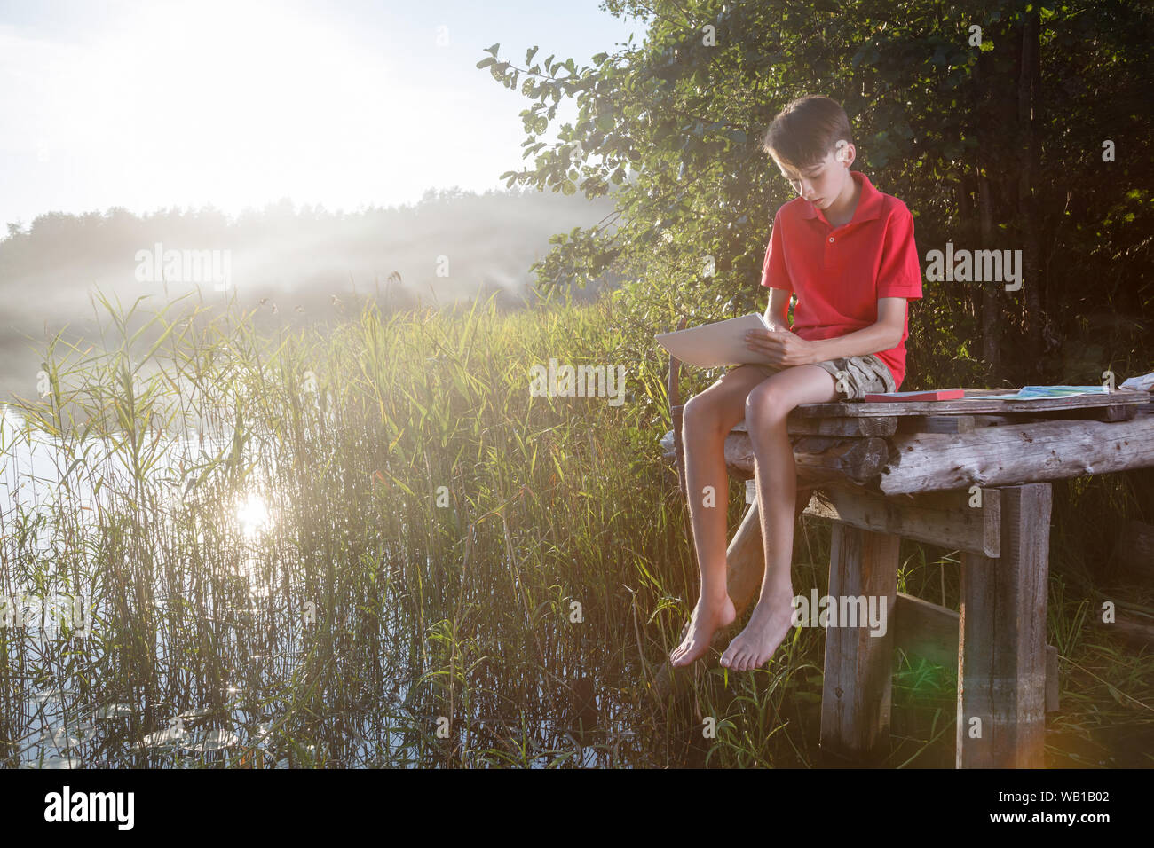 Adolescente ragazzo seduto su di un molo in legno dalla foresta lago di disegno con le matite di colore pastello Foto Stock