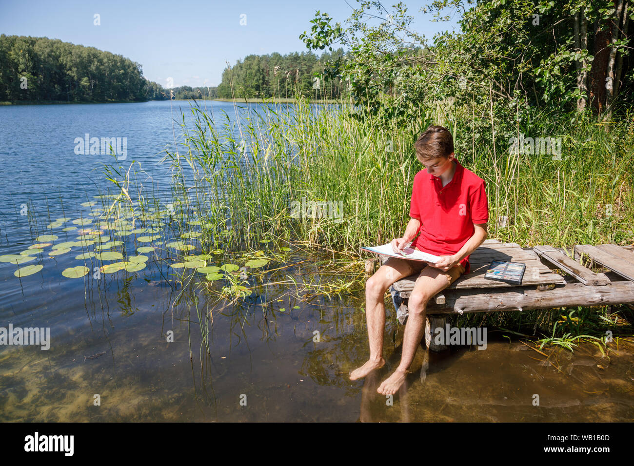 Adolescente ragazzo seduto su di un molo in legno dalla foresta lago di disegno con le matite di colore pastello Foto Stock
