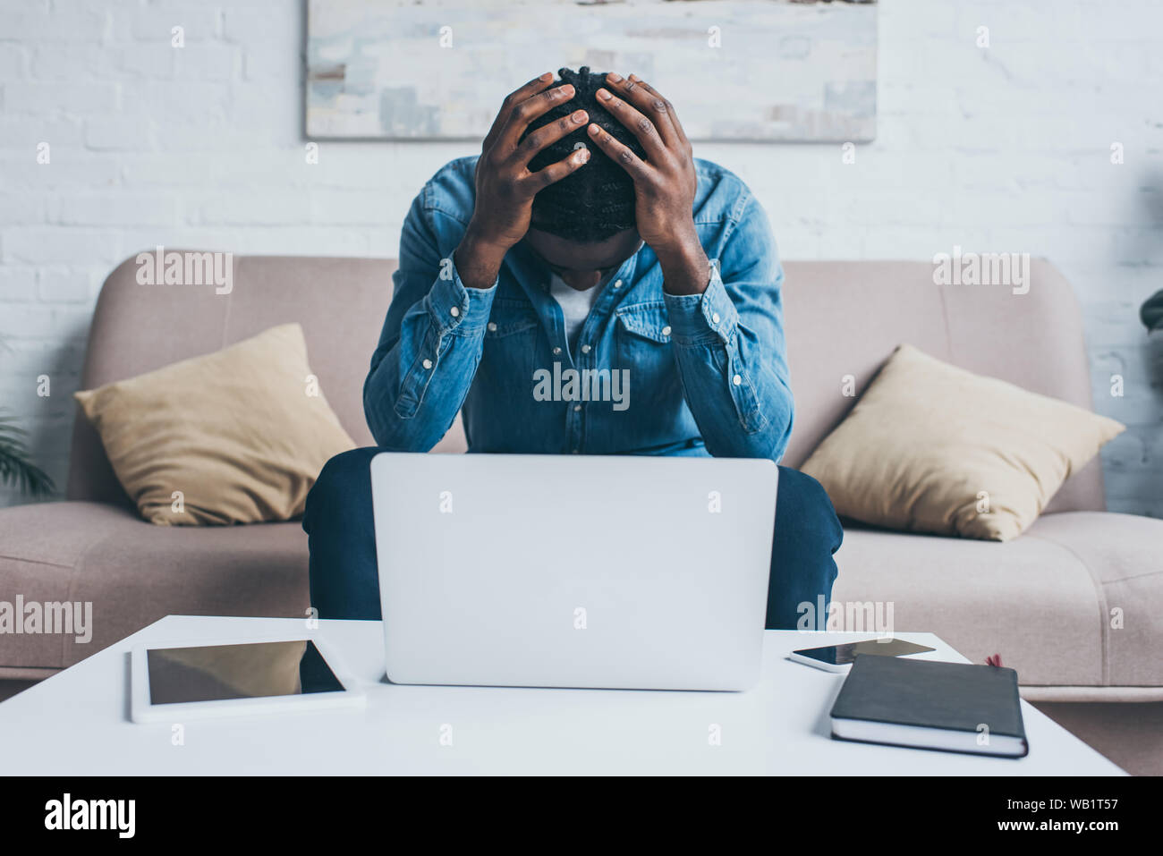 Stanco americano africano l uomo che soffre di mal di testa mentre è seduto vicino a tavola con i gadget Foto Stock