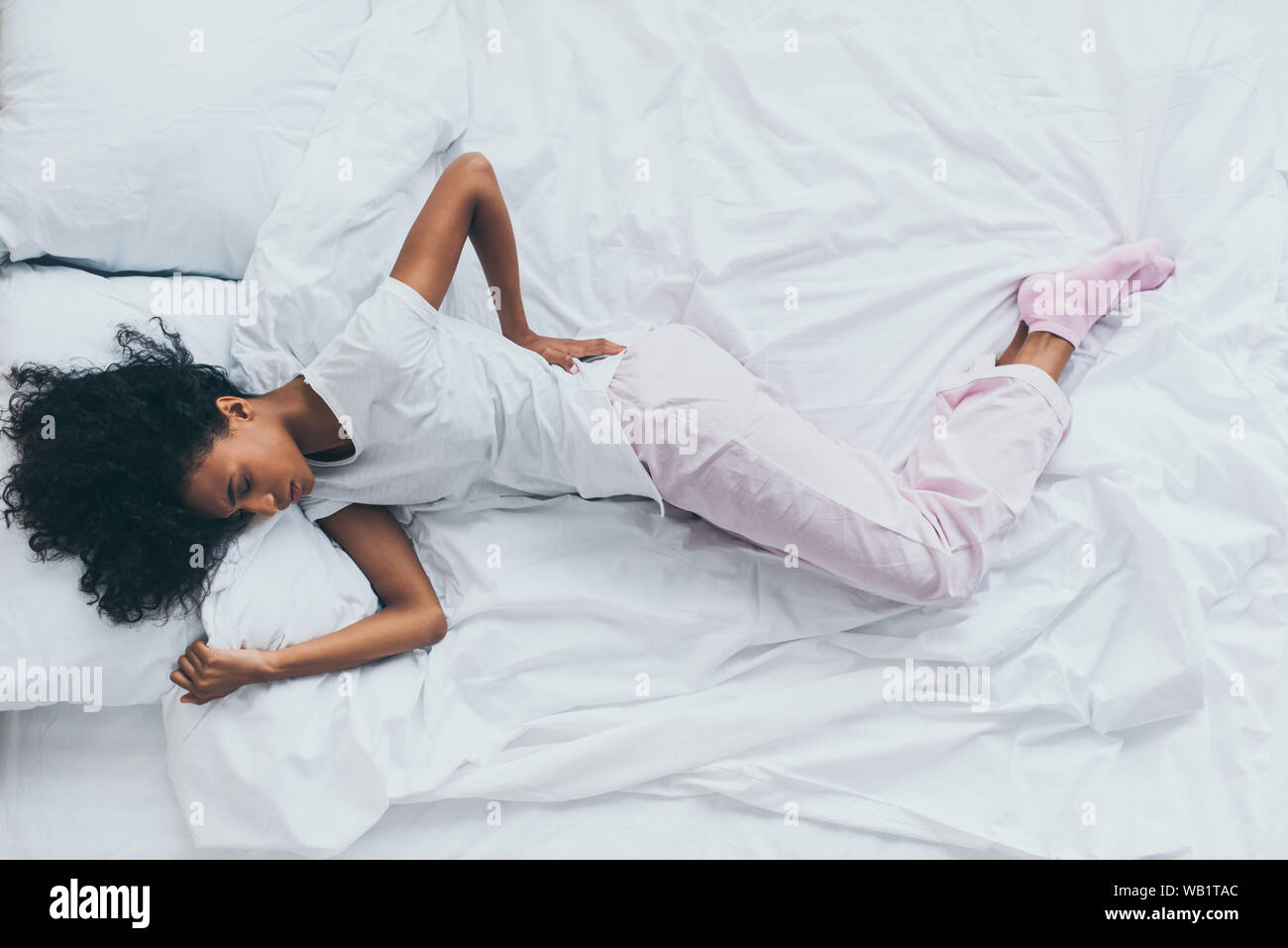 Vista superiore dell'African American donna che soffre di dolori alla schiena mentre giaceva sulla biancheria da letto bianca Foto Stock