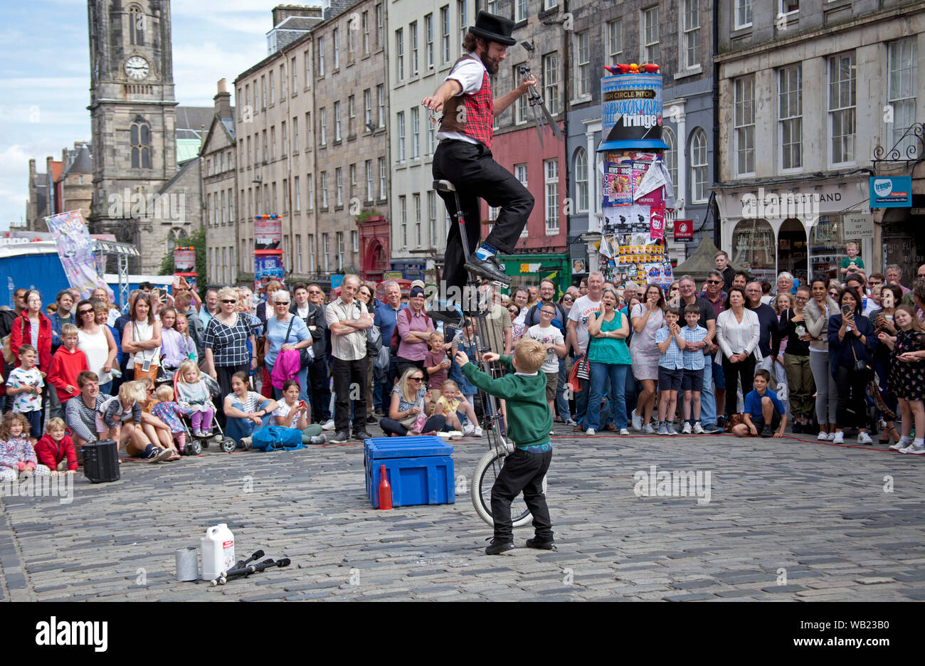 Il Royal Mile di Edimburgo, Scozia, Regno Unito. Il 23 agosto 2019. Street attore Patrick giocoliere e monociclo rider intrattiene il pubblico sulla High Street e teases sua giovane volontario con la promessa di denaro contante se egli salti per esso. Foto Stock