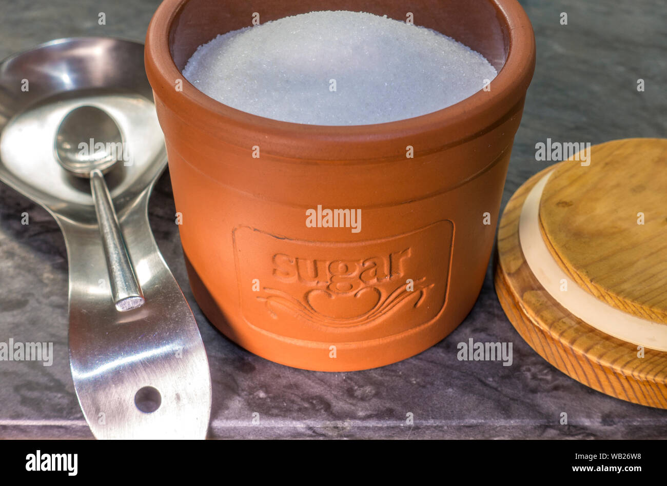 Chiudere il colpo di zucchero bianco in una terracotta contenitore di deposito, con il coperchio di legno e un cucchiaino di acciaio e il vassoio antigoccia accanto a. Foto Stock