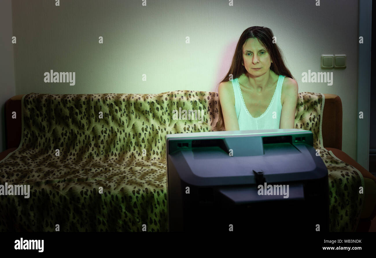 Una donna di mezza età, da soli, guarda la televisione, guardando triste, seduto su un divano. Lei guarda un triste film o ascolta la cattiva notizia che fa di lei un littl Foto Stock