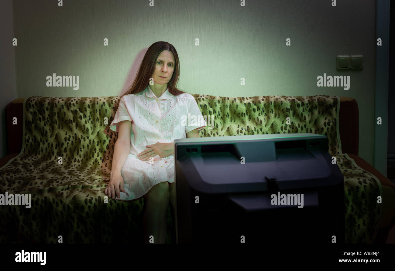 Una donna di mezza età, da soli, guarda la televisione, guardando triste, seduto su un divano. Lei guarda un triste film o ascolta la cattiva notizia che fa di lei un littl Foto Stock