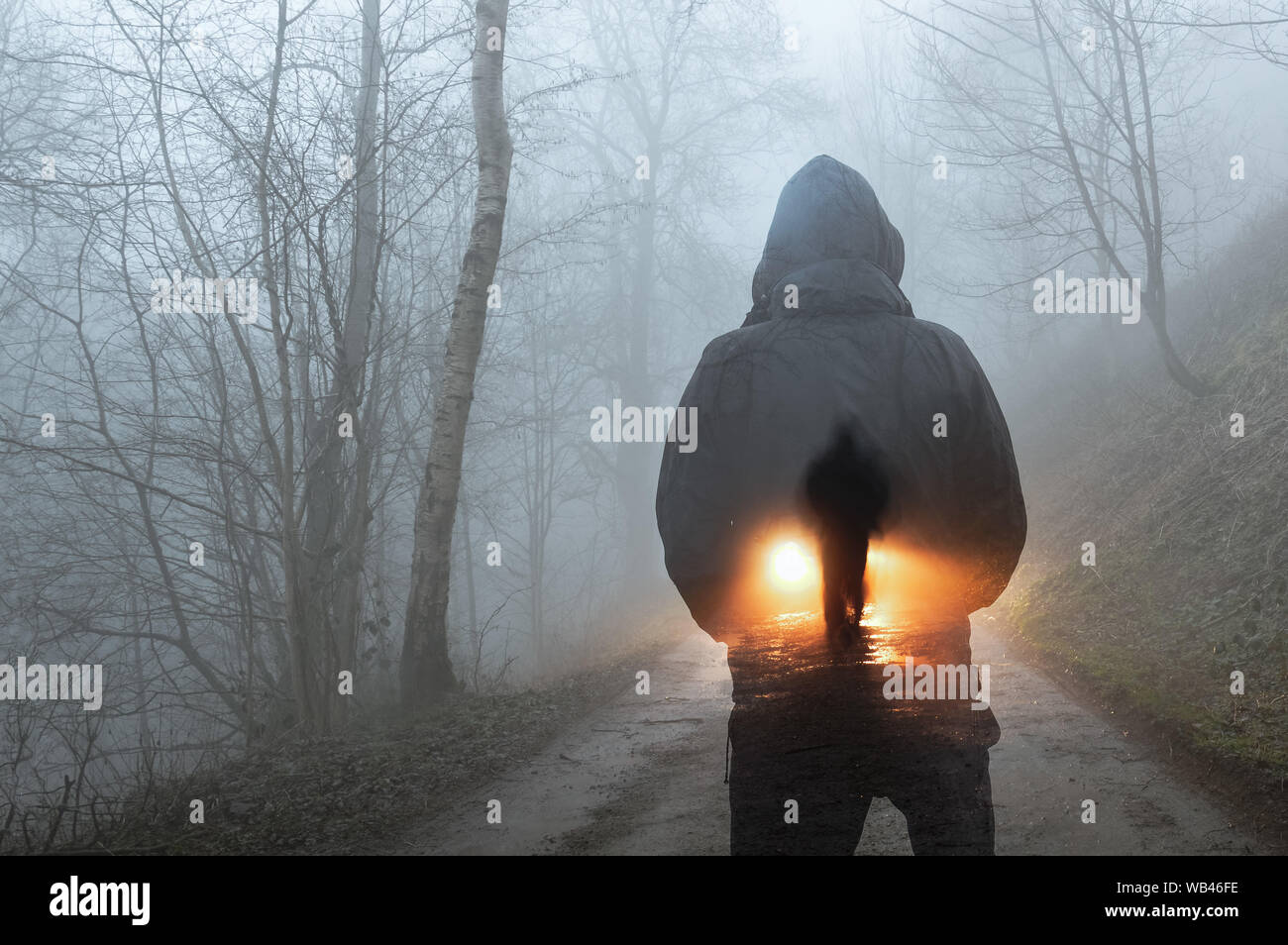 Una doppia esposizione concetto. Una figura con cappuccio guardando una silhouette di un uomo nella parte anteriore dei fari delle auto. In una foresta Spooky via in una nebbiosa inverni ev Foto Stock