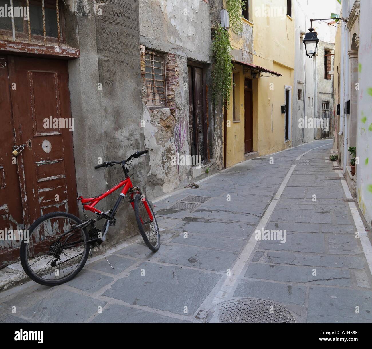 Una bicicletta rossa si appoggia contro una porta di legno in un lungo vialetto nella città vecchia di Rethymno, Creta. I muri in pietra e una linea che conduce nella distanza. Foto Stock