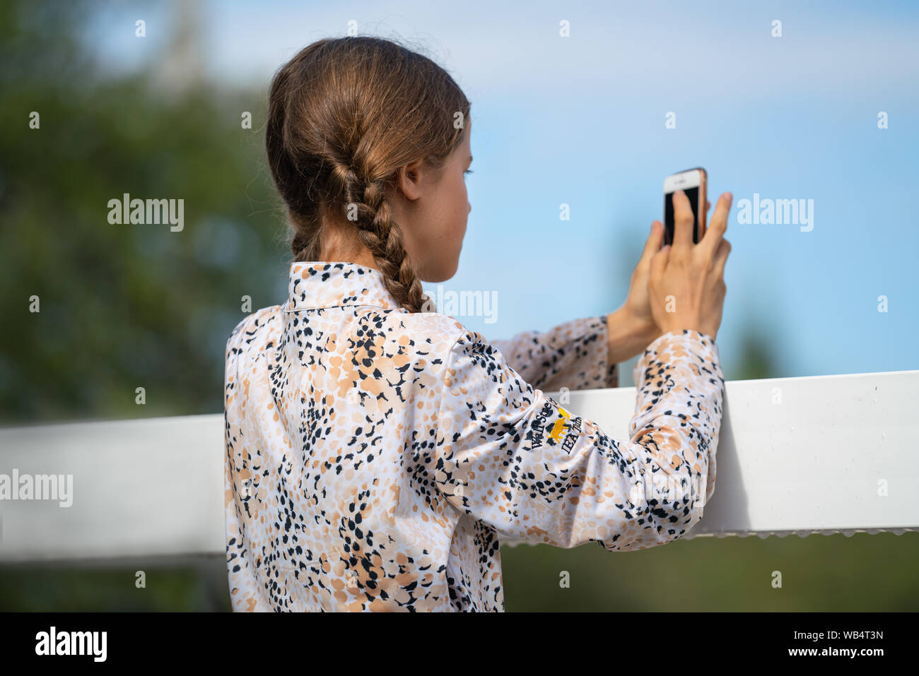 Una giovane ragazza caucasica con trecce è di scattare una foto a un evento equestre fuori con un telefono cellulare Foto Stock