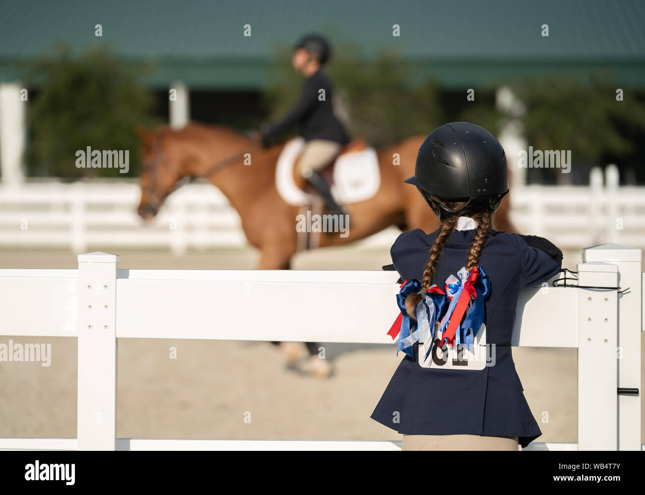 Giovane ragazza equestre con trecce è in piedi vicino al recinto della Mostra l'anello e la visione di un altro concorrente equestre Foto Stock