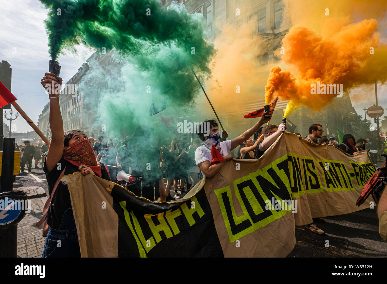 Londra, Regno Unito. 24 agosto 2019. Anti-fascisti attesa razzi di fumo come essi marzo per opporsi a una protesta presso la BBC da Tommy Robinson sostenitori che affermano che egli è in carcere per il giornalismo. Egli è stato condannato a 9 mesi per 3 reati al di fuori di Leeds Crown Court che potrebbe aver causato il crollo di una toelettatura pista di prova e ha precedenti condanne per violenza, finanziari e frodi in materia di immigrazione, del possesso di droga e di ordine pubblico reati. La polizia ha mantenuto la due gruppi separati. Robinson sostenitori sono stati successivamente unite dai dimostranti da Trafalgar Square, e un gruppo di grandi dimensioni dal supporto fino al razzismo è venuto per unire Antifa. Foto Stock