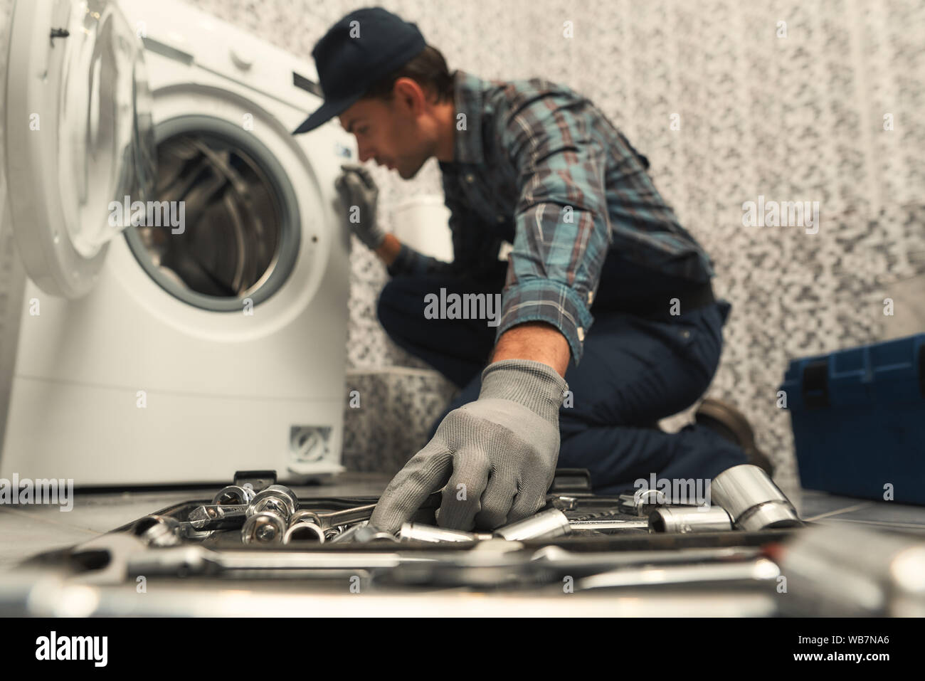 Ritratto di giovane tuttofare in uniforme seduto accanto a rotto la lavatrice e il tentativo di risolvere il problema. Inquadratura orizzontale. Vista laterale. Messa a fuoco selettiva su un lato e la casella degli strumenti Foto Stock