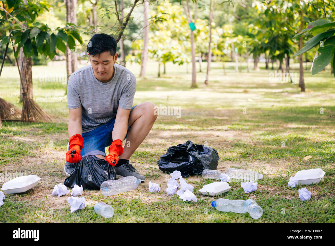 Giovane uomo asiatico indossando guanti di colore arancione e il cestino di raccolta nel sacchetto di immondizia nel parco. Salvare la terra e le preoccupazioni ambientali concetto Foto Stock