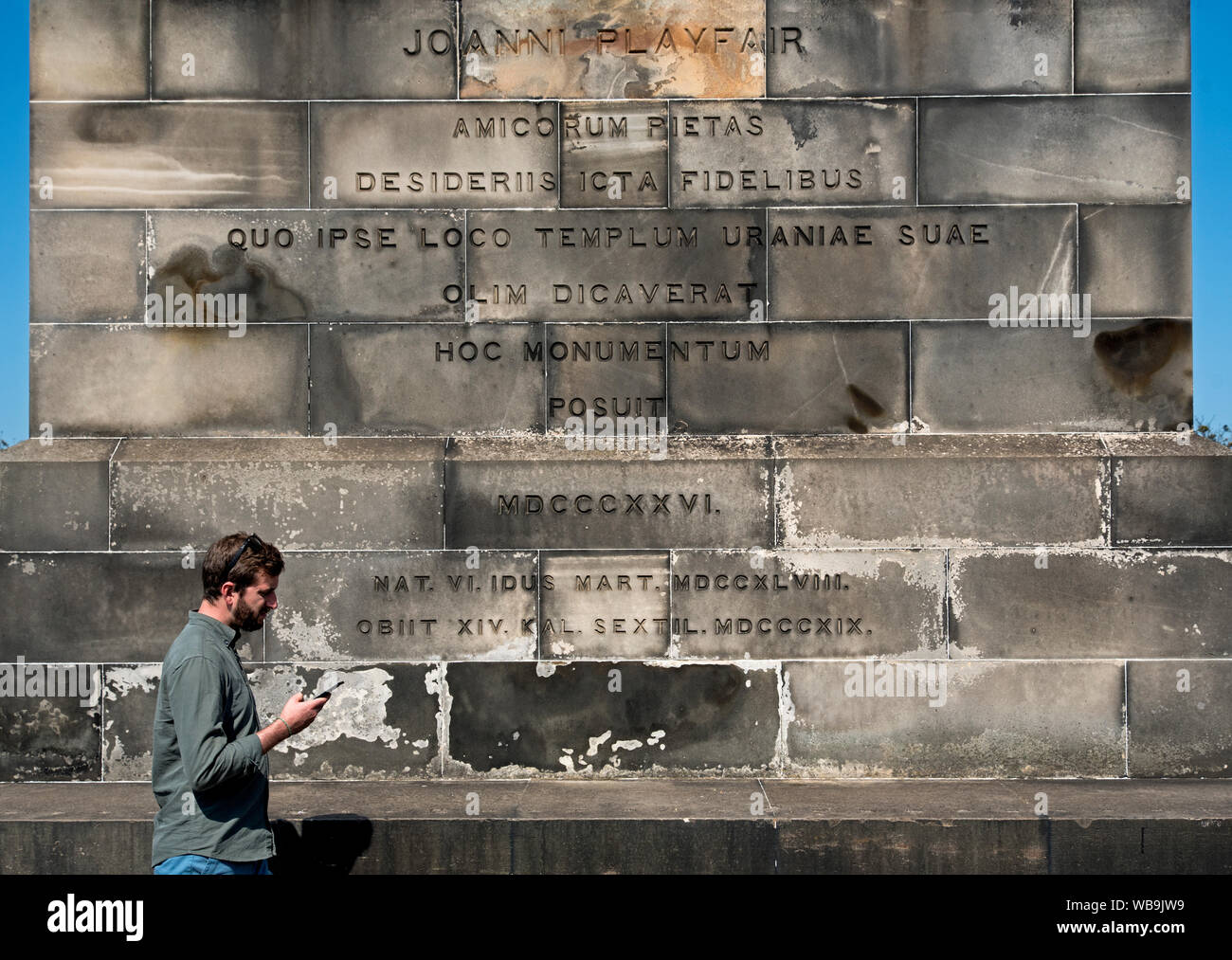 Un uomo che guarda il suo telefono cellulare passeggiate con la scritta in latino sul monumento a Giovanni Playfair (1748-1819) su Calton Hill, Edimburgo, Scozia, Regno Unito. Foto Stock