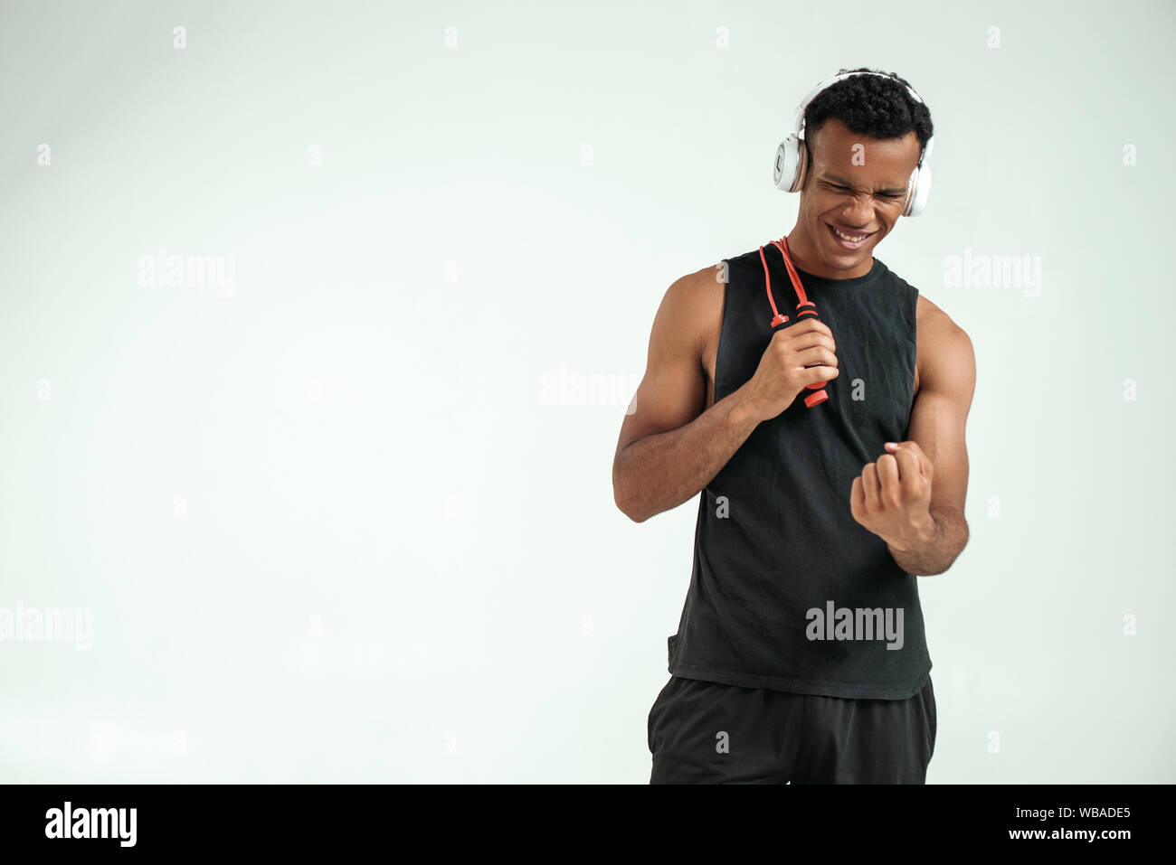 Non posso fare tutto. Allegro giovani afro-americano nelle cuffie tenendo un salto con la corda, sorridente e stringendo un pugno in piedi contro uno sfondo grigio. Attrezzature sportive. Motivazione Foto Stock