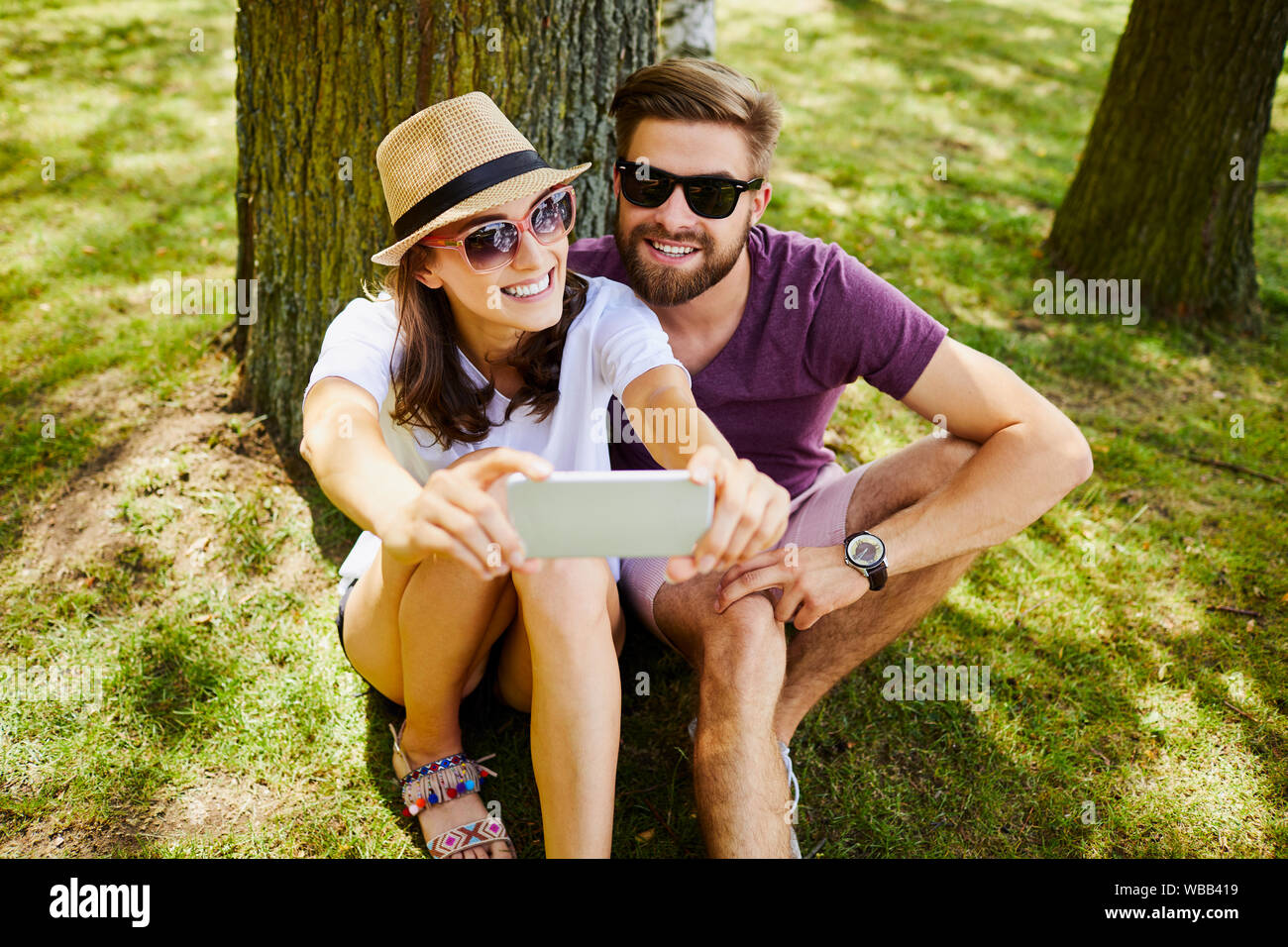 Coppia giovane tenendo selfie mentre è seduto di fronte ad un albero in un parco durante l'estate Foto Stock