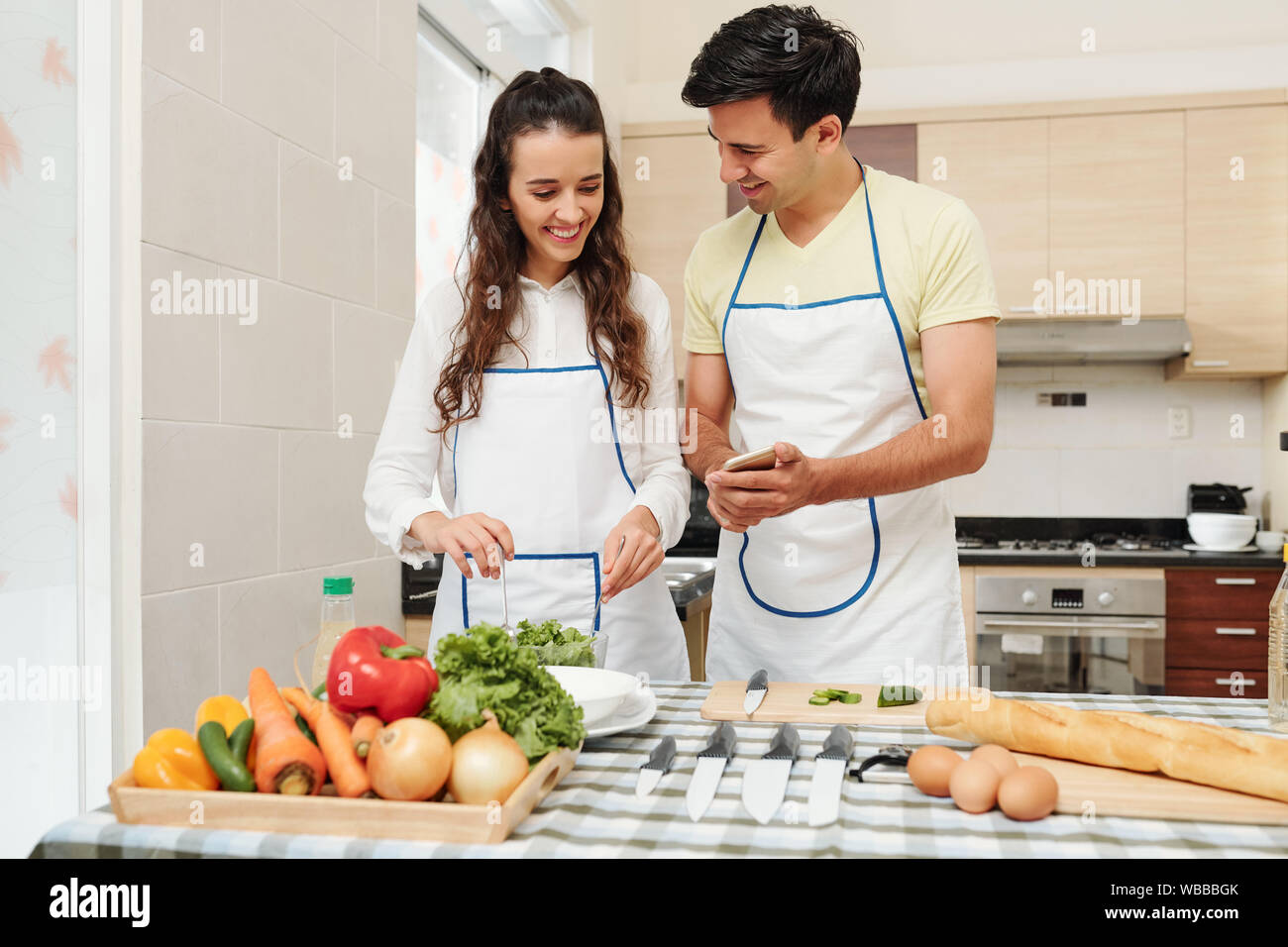 Allegro giovane uomo che mostra immagini divertenti sulla schermata dello smartphone alla ragazza che è la miscelazione di una sana insalata per la cena Foto Stock