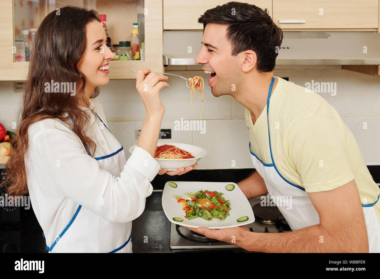 Felice giovane donna in amore alimentando il suo fidanzato con gli spaghetti quando sono in piedi in cucina dopo cena di cucina Foto Stock