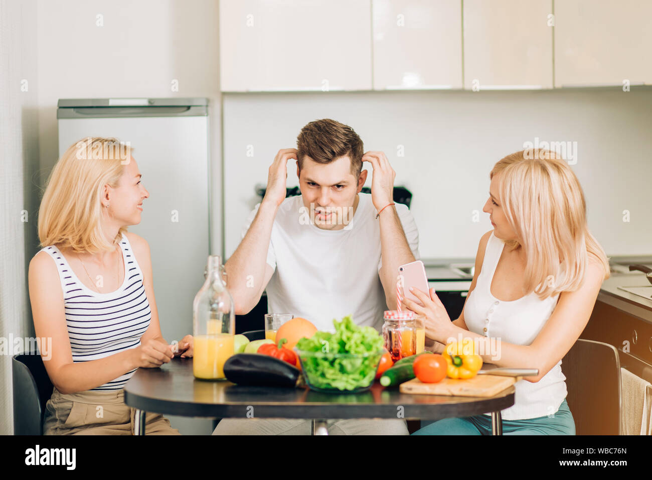Due belle caucasica bionde ascoltando il loro amico racconta la storia interessante. Tre amici seduti a tavola in cucina, potabile e frutta veget Foto Stock