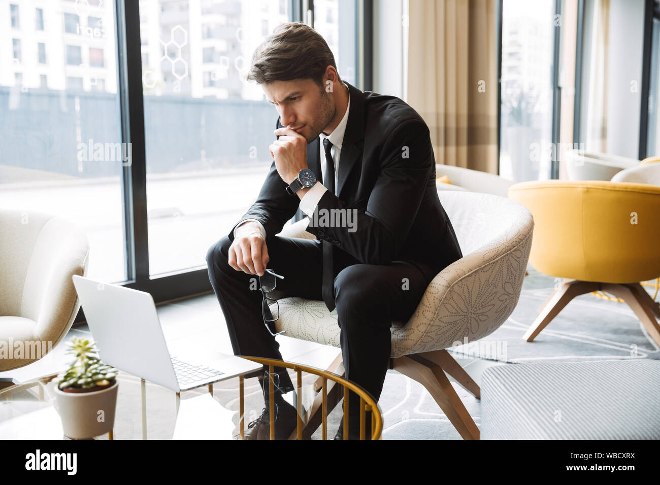 Foto di concentrato di imprenditore formale da indossare tuta nera utilizzando il computer portatile nella hall hotel Foto Stock