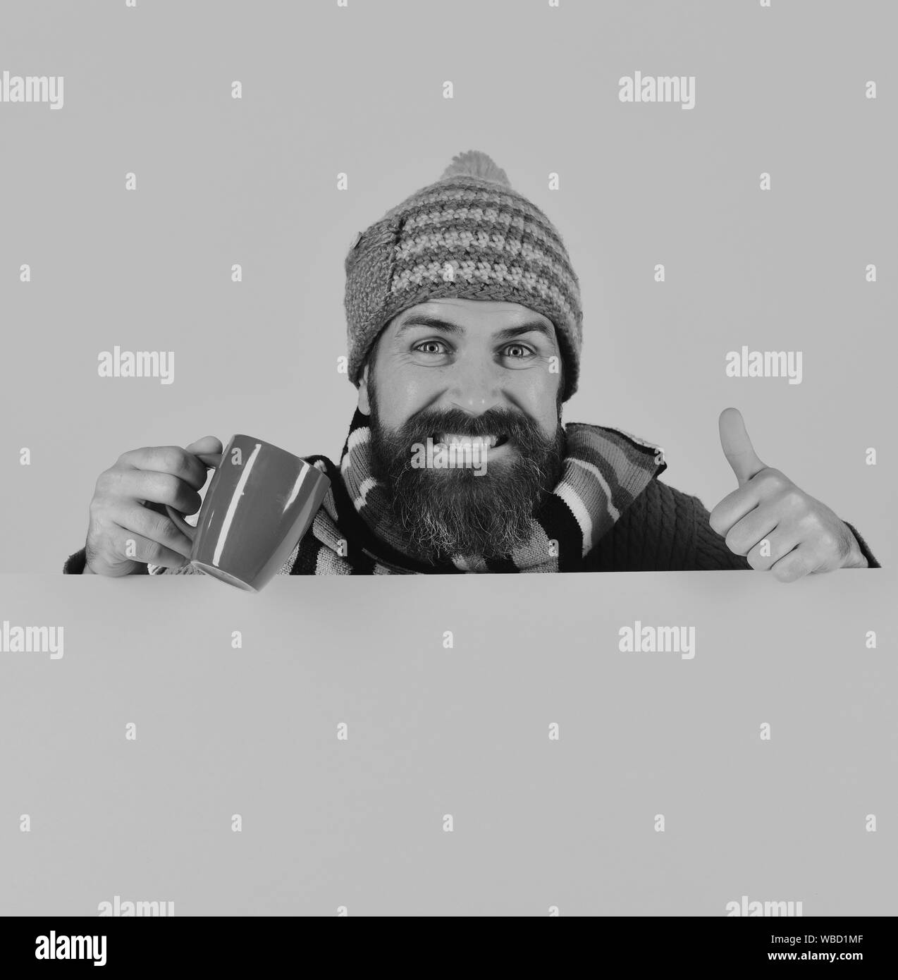Perfetto mattina autunnale e concetto di aroma. Il momento della colazione idea. Uomo in caldo hat detiene tazza marrone su sfondo rosa, copia dello spazio. Hipster con la barba e la faccia felice dispone di tazza di caffè mostrando pollice in alto Foto Stock