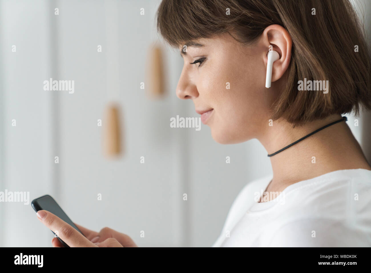 Immagine positiva di un giovane bella donna al chiuso in casa ascoltando la musica con gli auricolari bluetooth utilizzando il telefono cellulare. Foto Stock