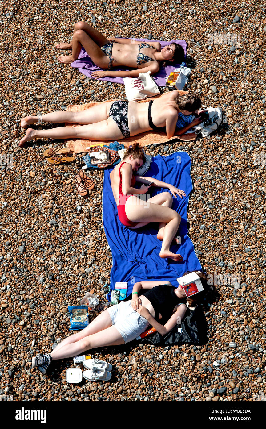 Quattro giovani donne rilassarsi leggendo, sms e prendere il sole sulla spiaggia di Brighton nell'ondata di caldo e di registrare le temperature elevate dell'estate 2019. Foto Stock