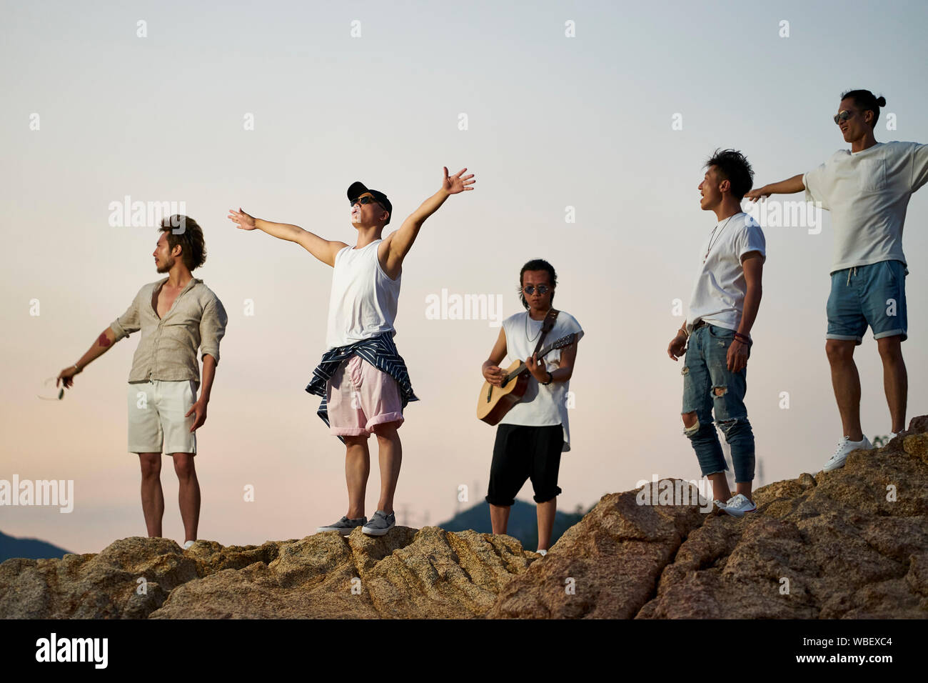 I membri di una rock band in piedi sulla parte superiore delle rocce in riva al mare a cantare a suonare la chitarra Foto Stock