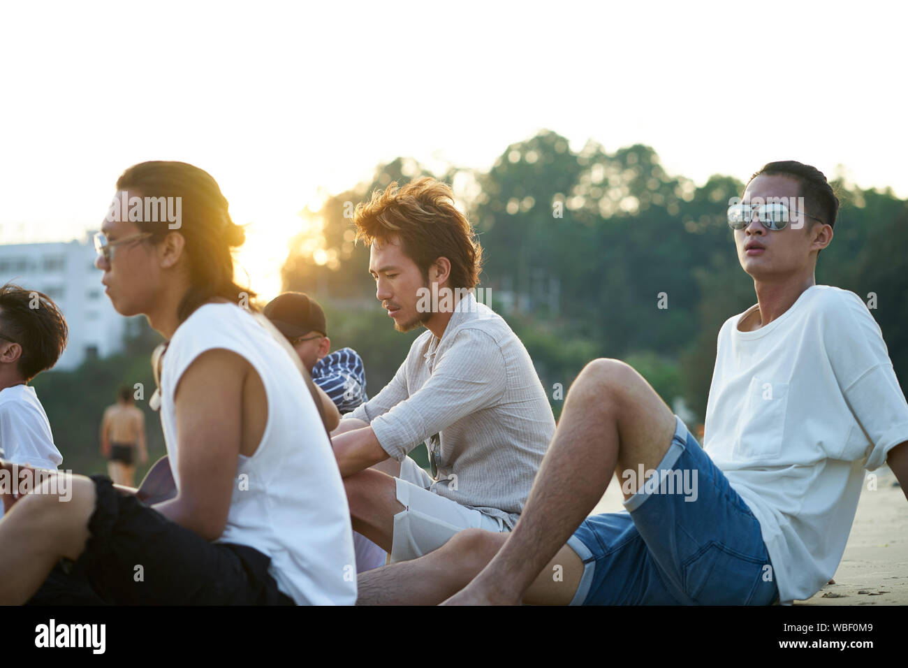 Il gruppo di cinque giovani uomini asiatici seduto sulla spiaggia di sabbia al tramonto Foto Stock
