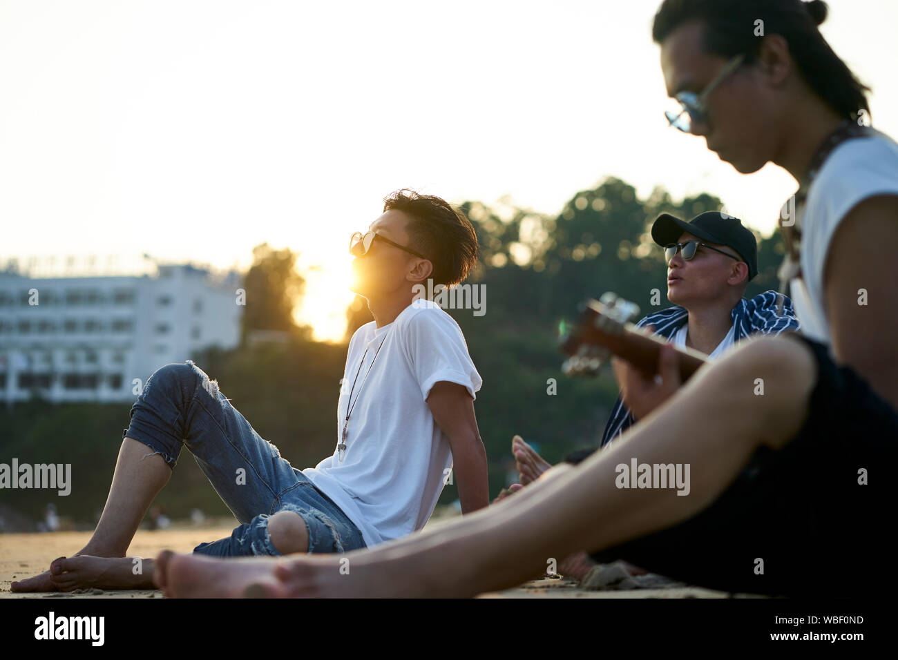 Tre giovani asiatici uomini adulti seduti sulla spiaggia di sabbia a suonare la chitarra al crepuscolo Foto Stock