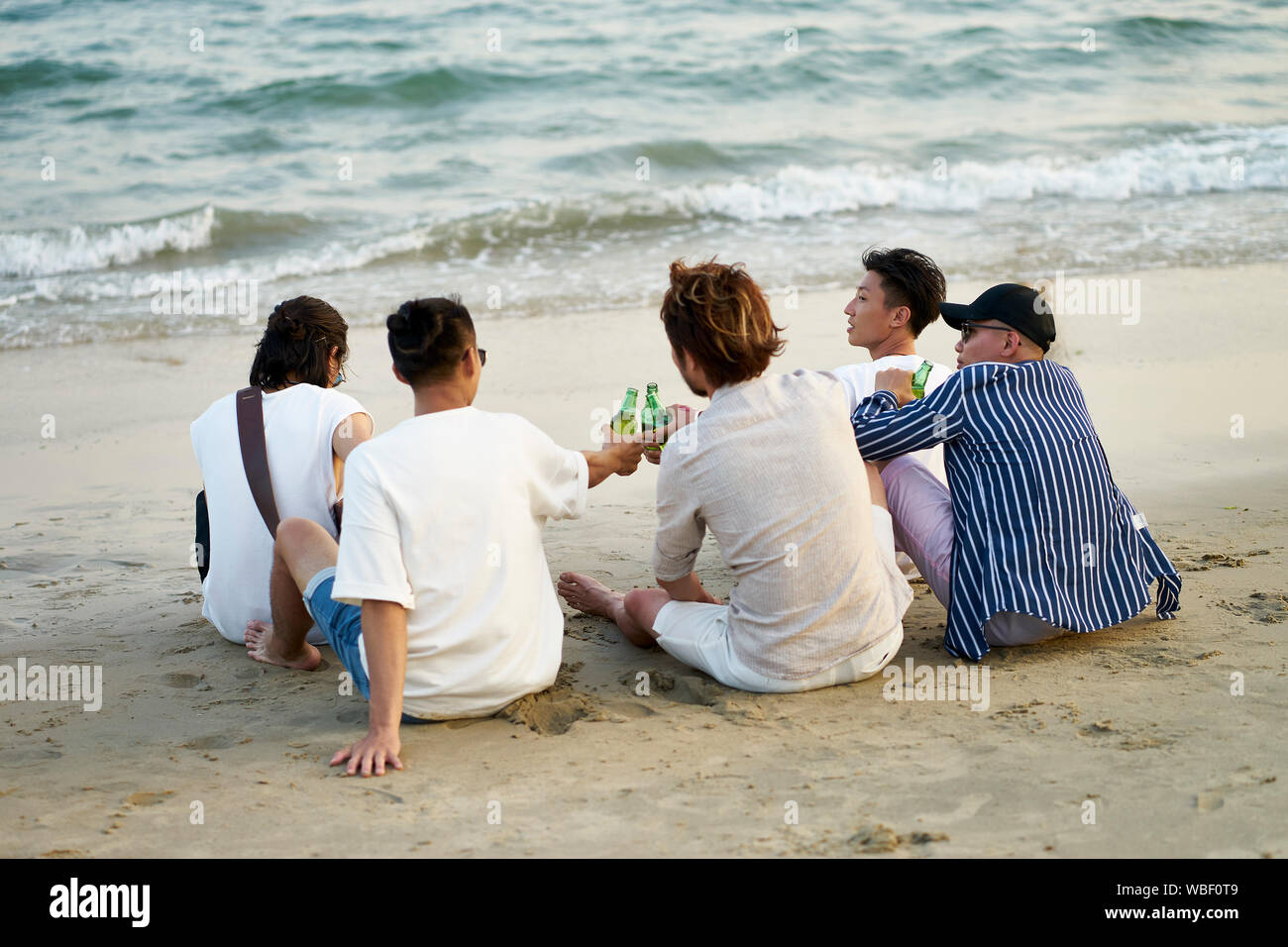 Un gruppo di giovani asiatici uomini adulti bere birra facendo clic su bottiglie tostare sulla spiaggia, vista posteriore Foto Stock