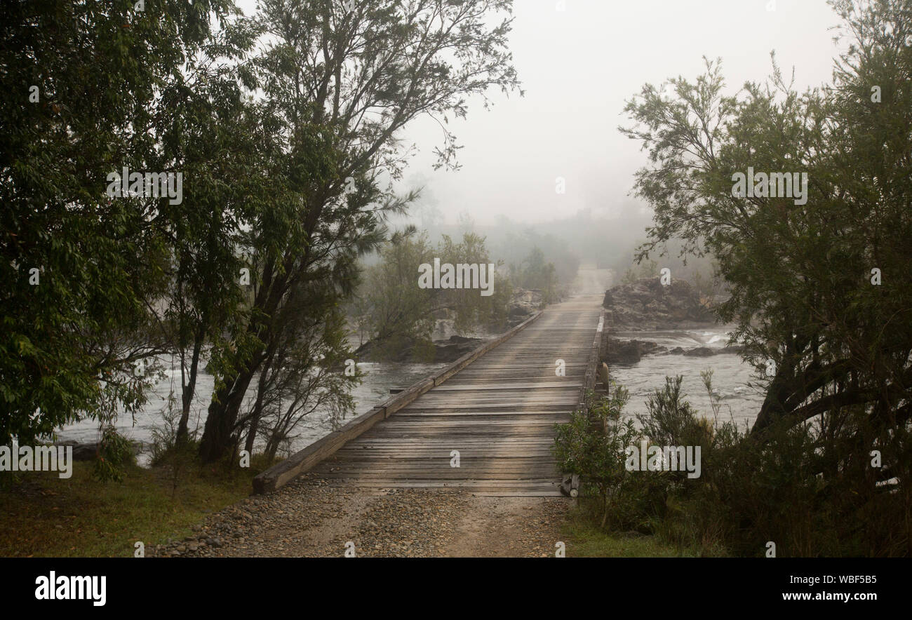 Lunga e stretta strada in legno ponte che attraversa il rock-disseminata Mann fiume orlate da alberi nativi e scomparire nella nebbia di mattina presto in NSW Australia Foto Stock